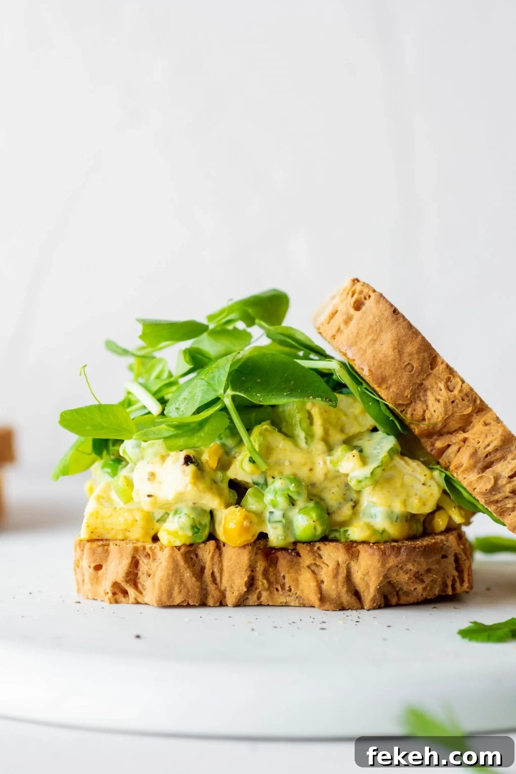 Side view of a curried chicken salad sandwich with some fresh greens and the top piece of bread angled off to the side. On a white background.
