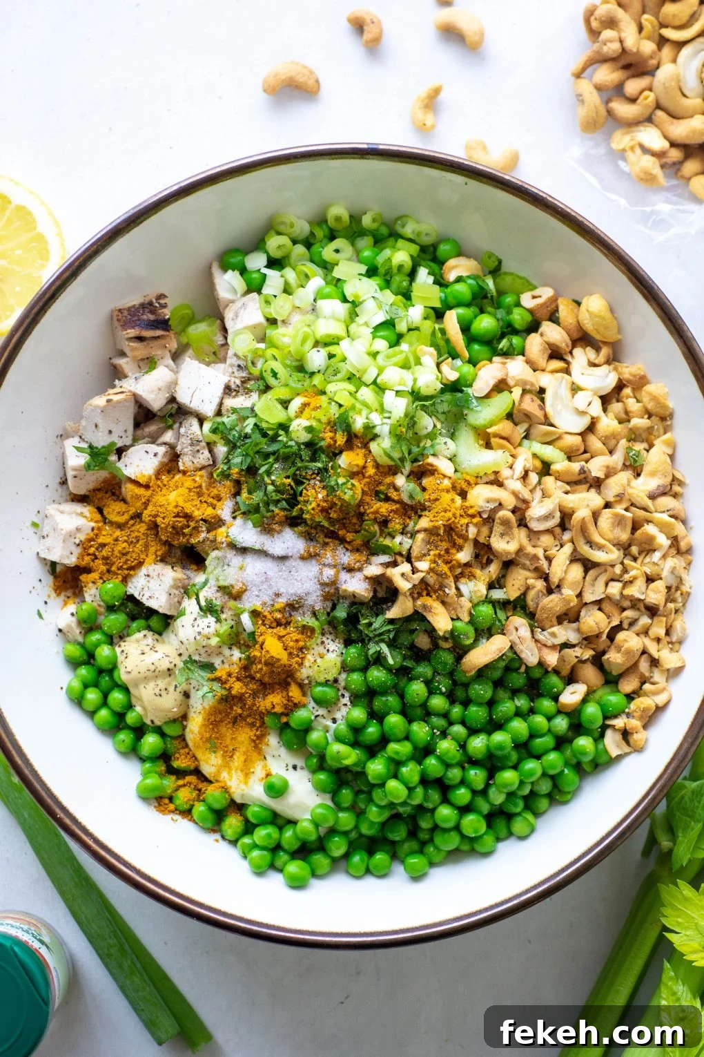 Overhead view of a bowl filled with curried chicken salad ingredients - chopped chicken, green peas, celery, green onions, cashews, mayo, and spices.