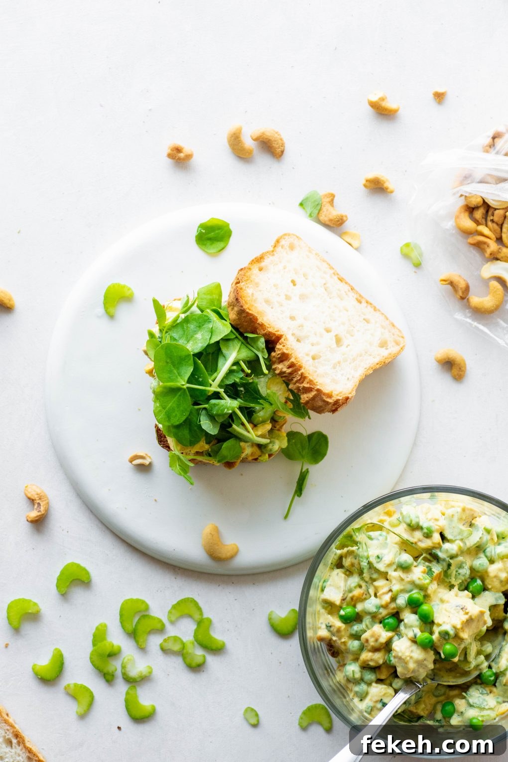 Overhead shot of an open faced chicken curry salad sandwich topped with salad greens on a white background. Surrounded by a bowl of chicken salad, some toasted cashews, and sliced celery.