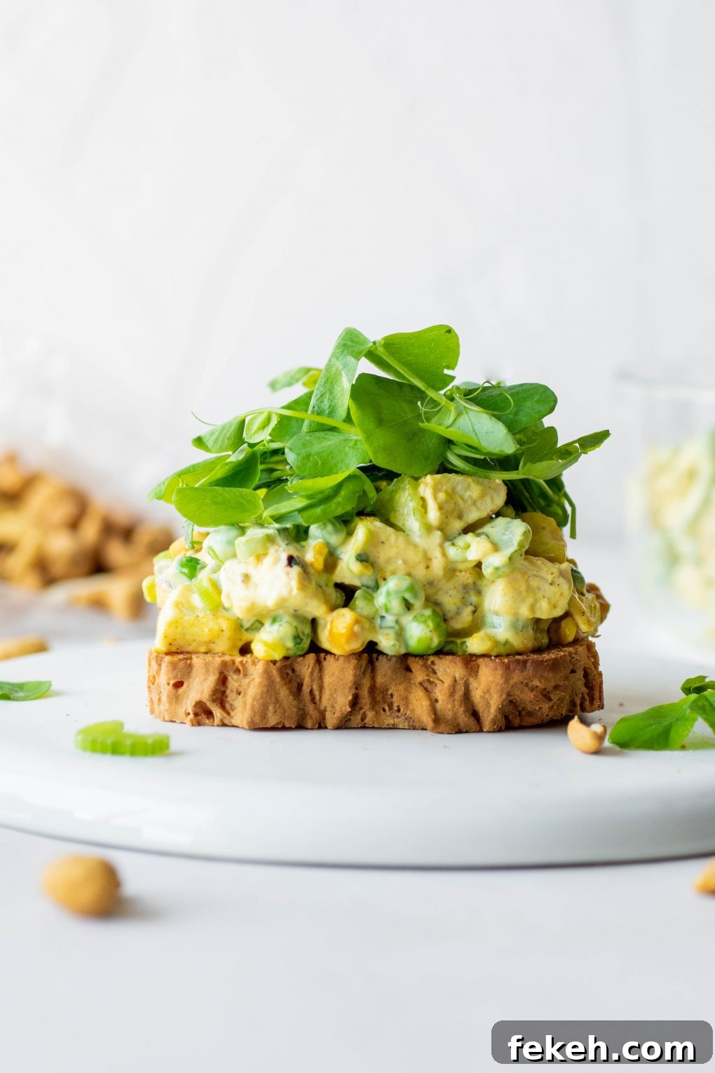 Close up side view of an open faced chicken curry salad sandwich with bright greens on a white background.