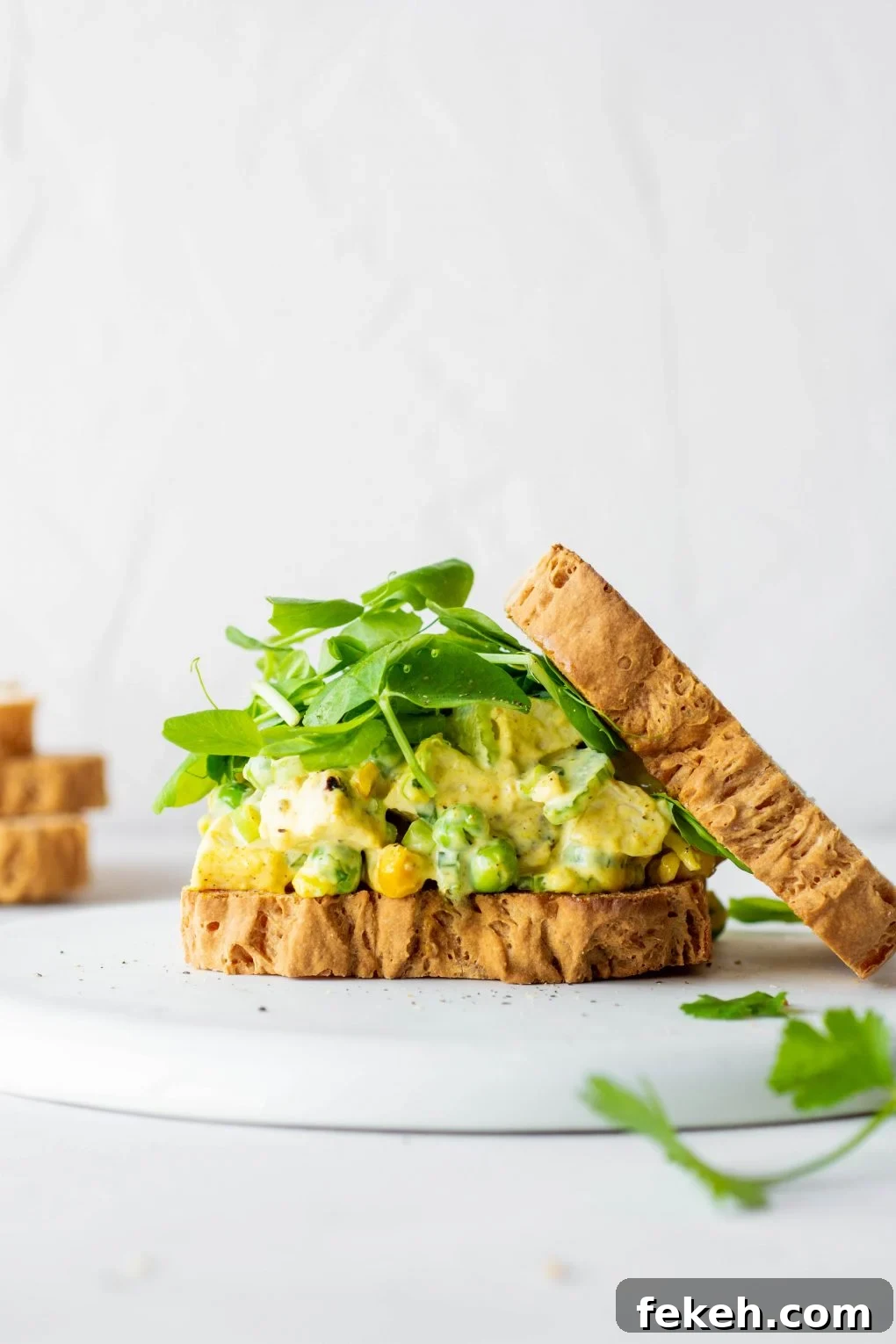 Side view of a curried chicken salad sandwich with some fresh greens and the top piece of bread angled off to the side. On a white background.