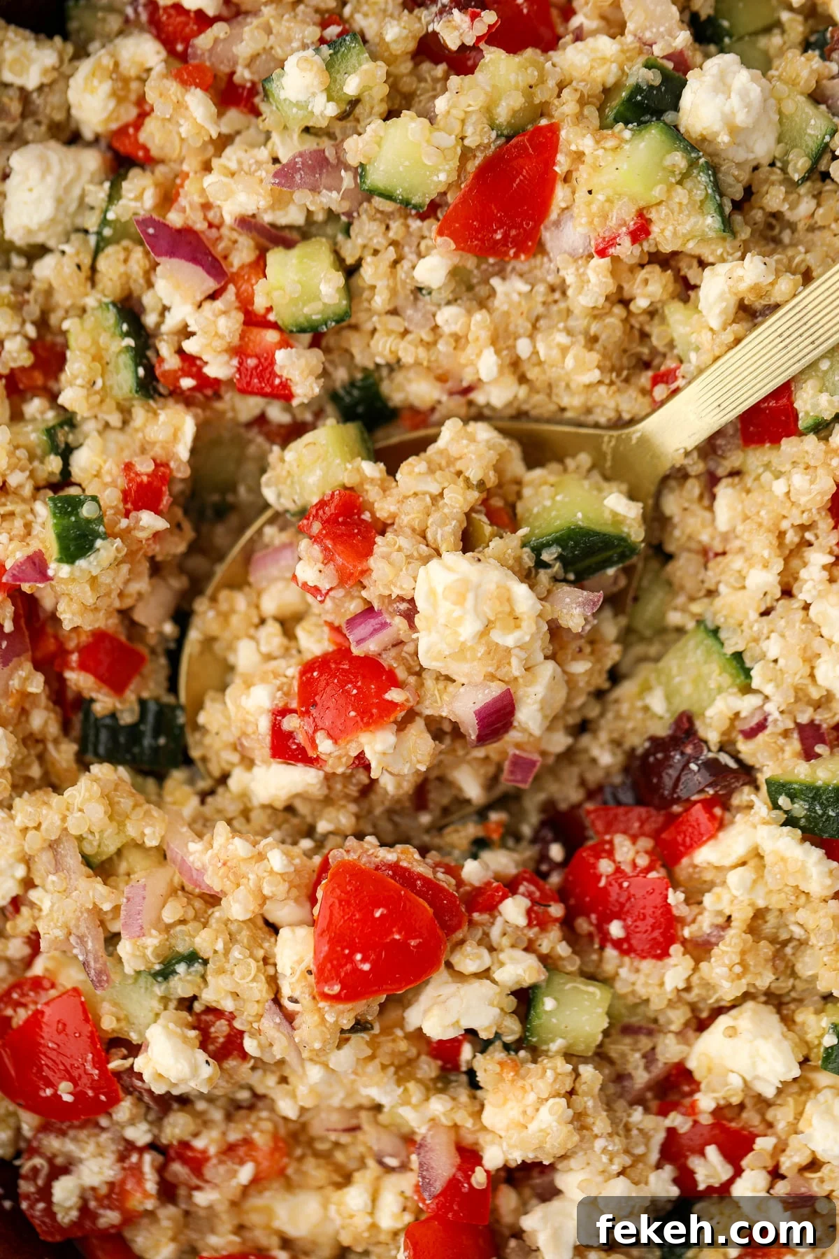 A spoon scooping into quinoa salad featuring red pepper, feta, and cucumbers.