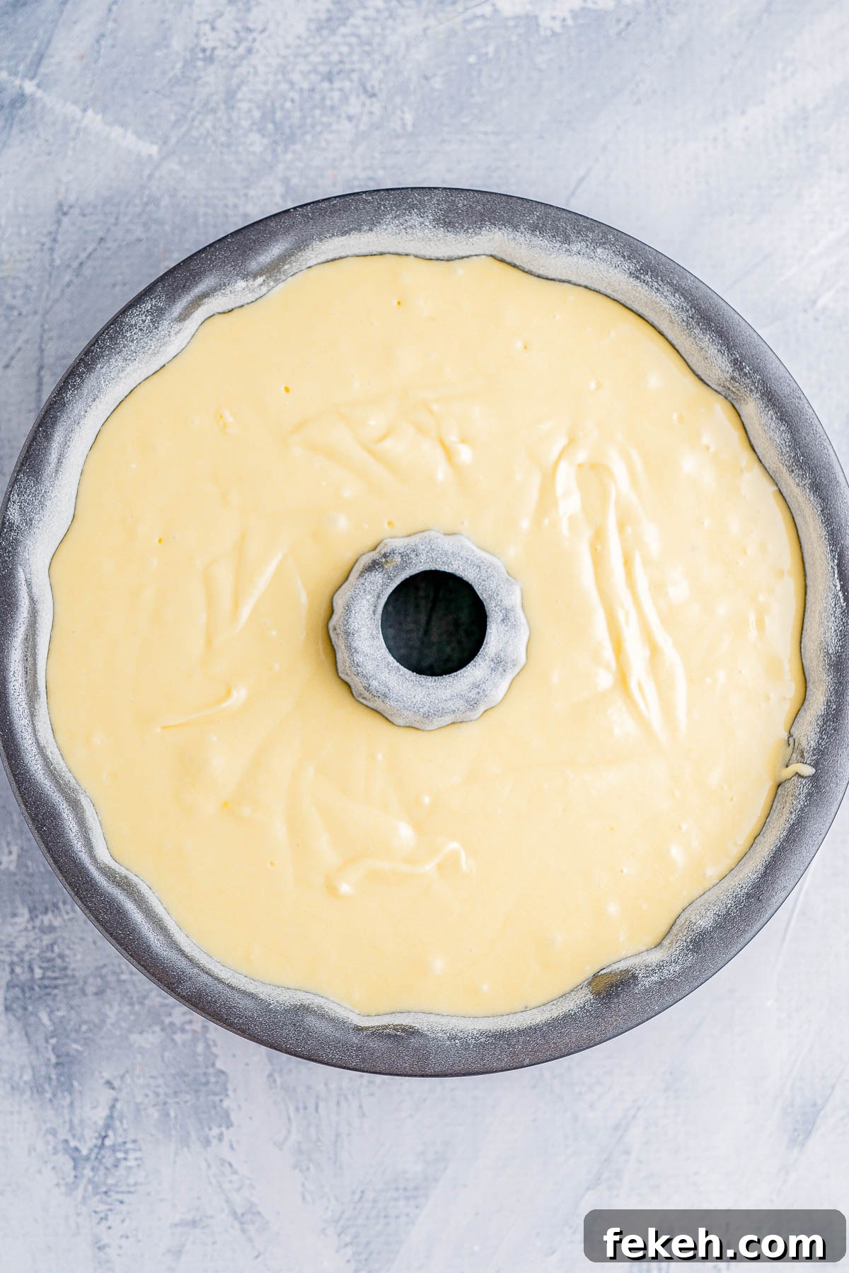 Classic Cream Cheese Pound Cake Recipe for Moist, Tender Slices 11 A round, gray bundt pan filled with yellow cake batter, ready for baking, on a light textured surface.