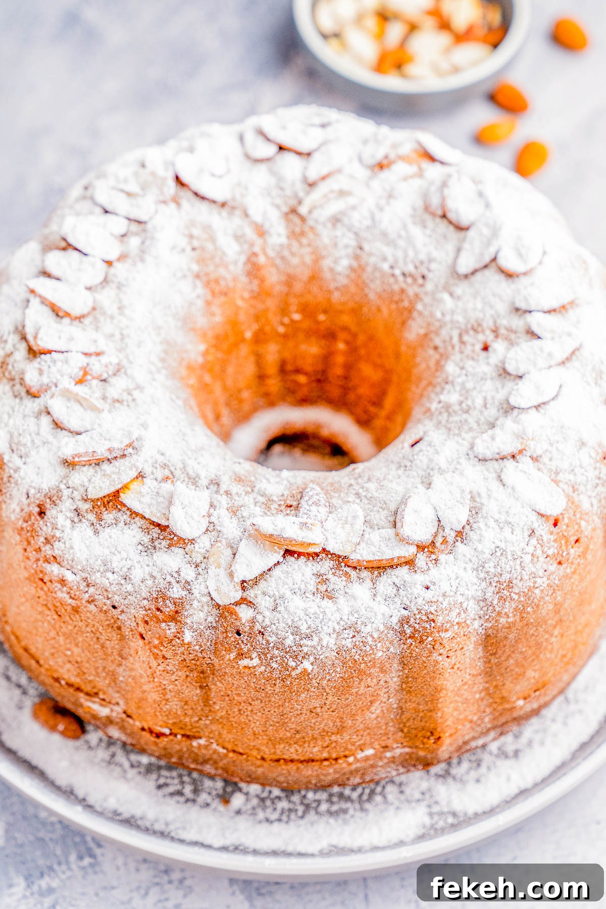 Classic Cream Cheese Pound Cake Recipe for Moist, Tender Slices 14 Bundt cake topped with powdered sugar and almond slices on a plate, with a small bowl of almond slices in the background.