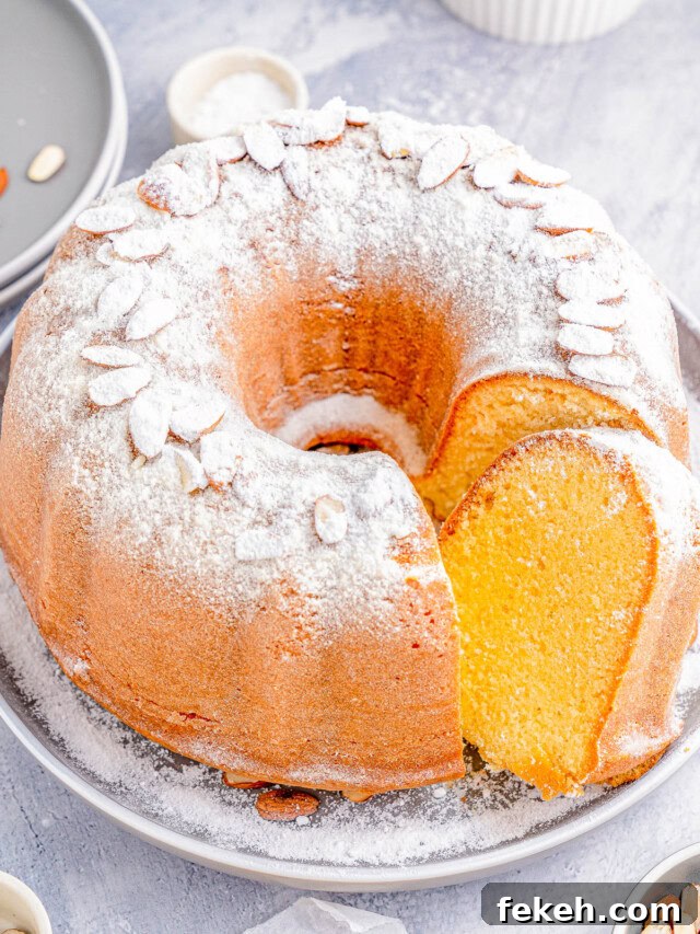 Classic Cream Cheese Pound Cake Recipe for Moist, Tender Slices 22 Bundt cake dusted with powdered sugar and topped with almond slices on a gray plate, featuring a single slice cut out; surrounded by scattered almonds and small dishes.