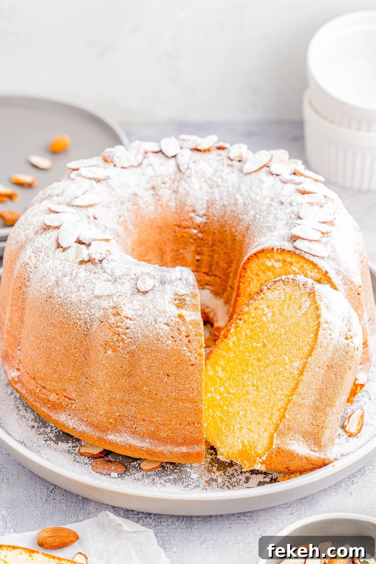Classic Cream Cheese Pound Cake Recipe for Moist, Tender Slices 7 A Bundt cake on a plate, dusted with powdered sugar and topped with sliced almonds. A slice is cut from the cake. Plates and dishes are in the background.