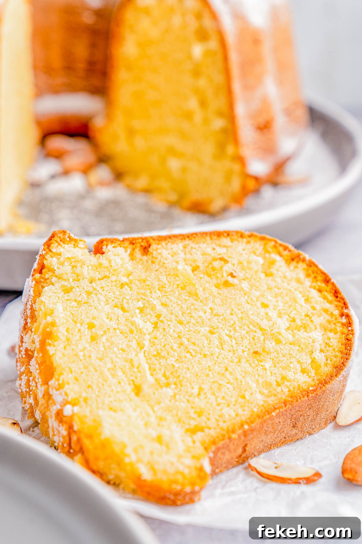 Classic Cream Cheese Pound Cake Recipe for Moist, Tender Slices 8 A close-up of a slice of a Bundt cake on a plate, with a dusting of powdered sugar and almond slices nearby.