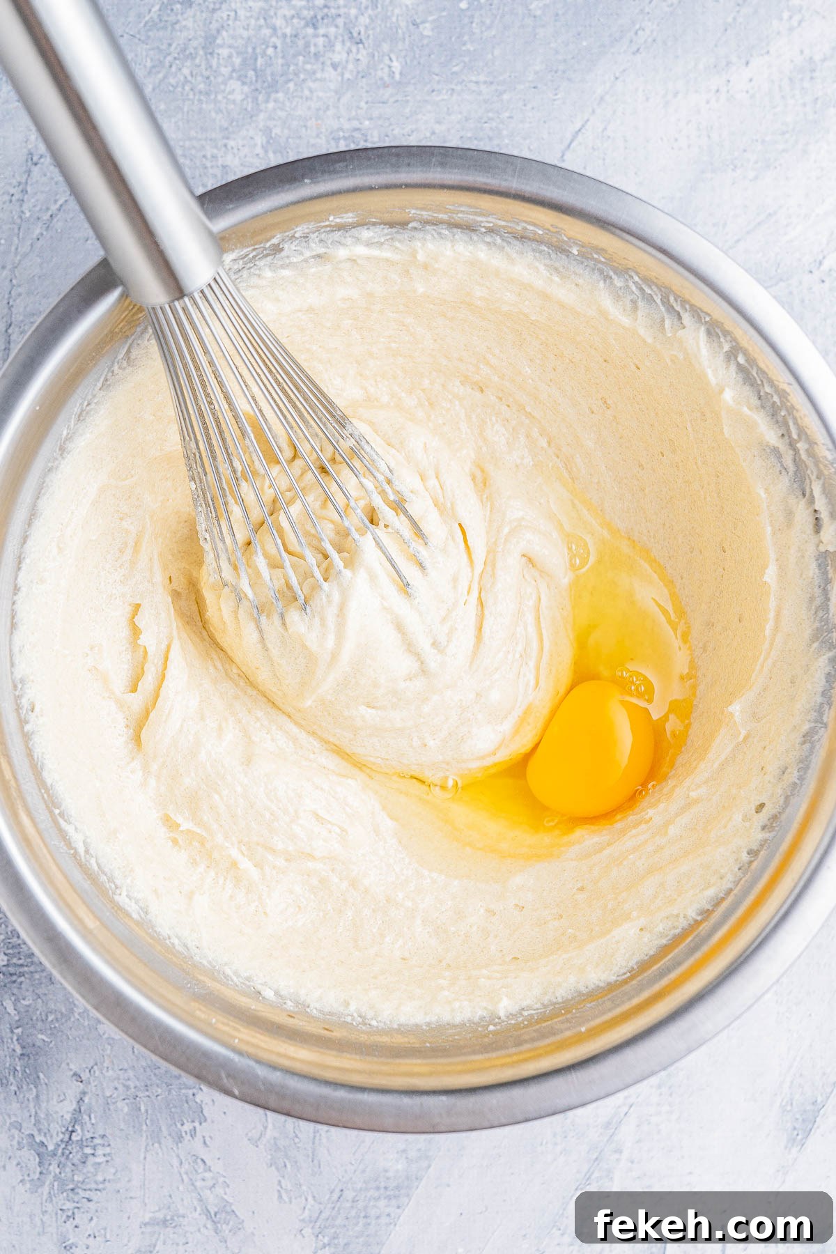 Classic Cream Cheese Pound Cake Recipe for Moist, Tender Slices 9 A whisk mixing a yellow egg into a creamy batter in a stainless steel bowl on a textured surface.