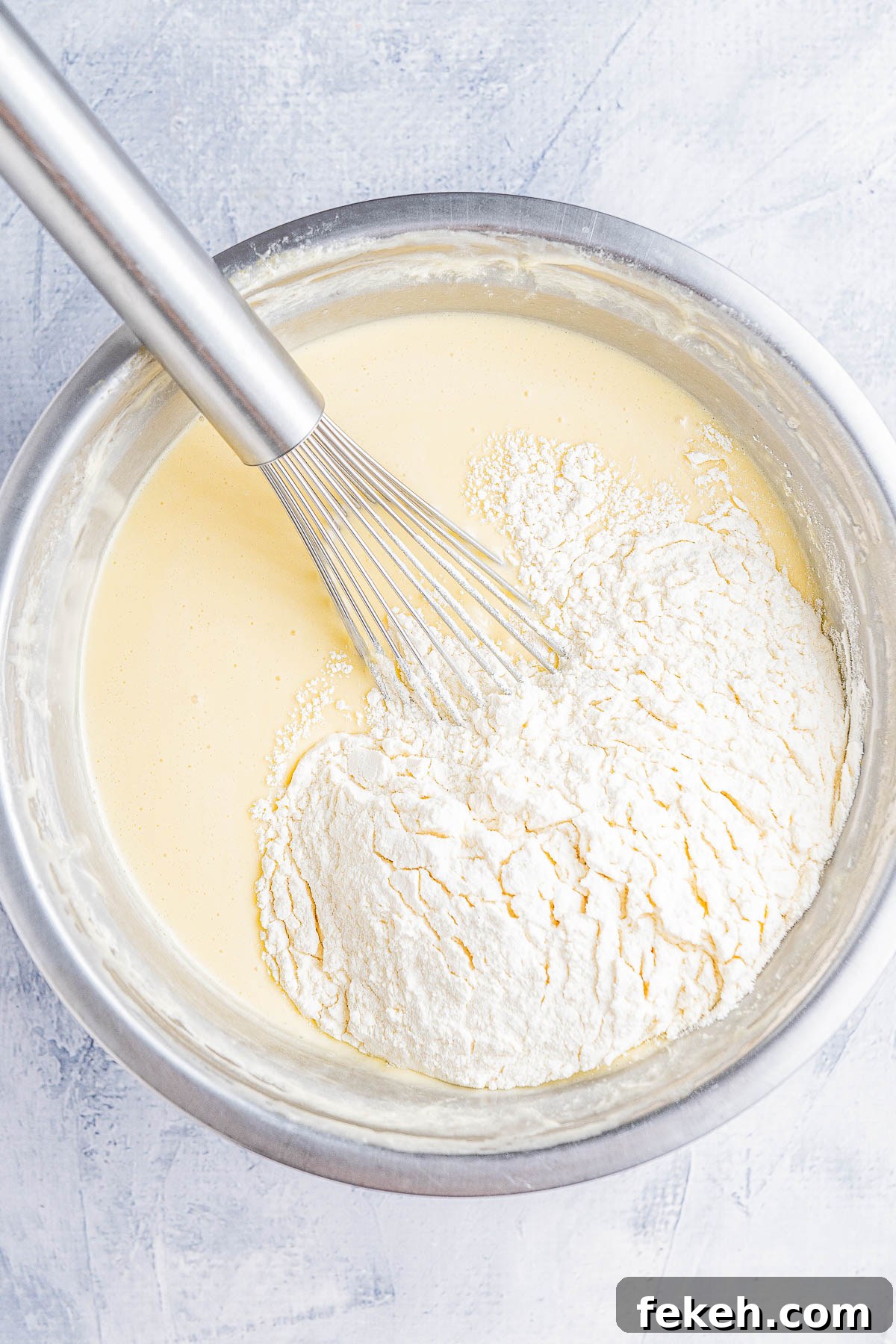 Classic Cream Cheese Pound Cake Recipe for Moist, Tender Slices 10 A metal bowl with flour being whisked into a creamy batter using a metal whisk, placed on a light gray textured surface.