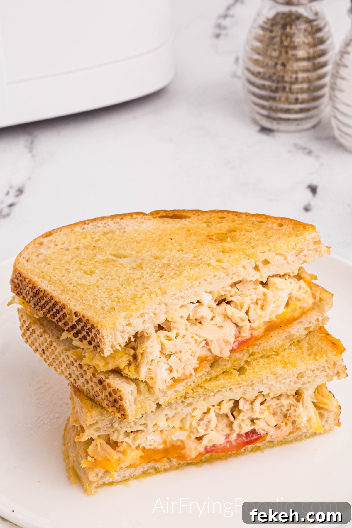 Crispy Air Fryer Tuna Melt Sandwich Recipe 2 Tuna melt on golden Texas toast cooked in the air fryer with tomato and cheese cut in half and served on a round white plate. A small portion of the air fryer base is in the top left of the picture, and a sale and pepper shaker is in the top right.
