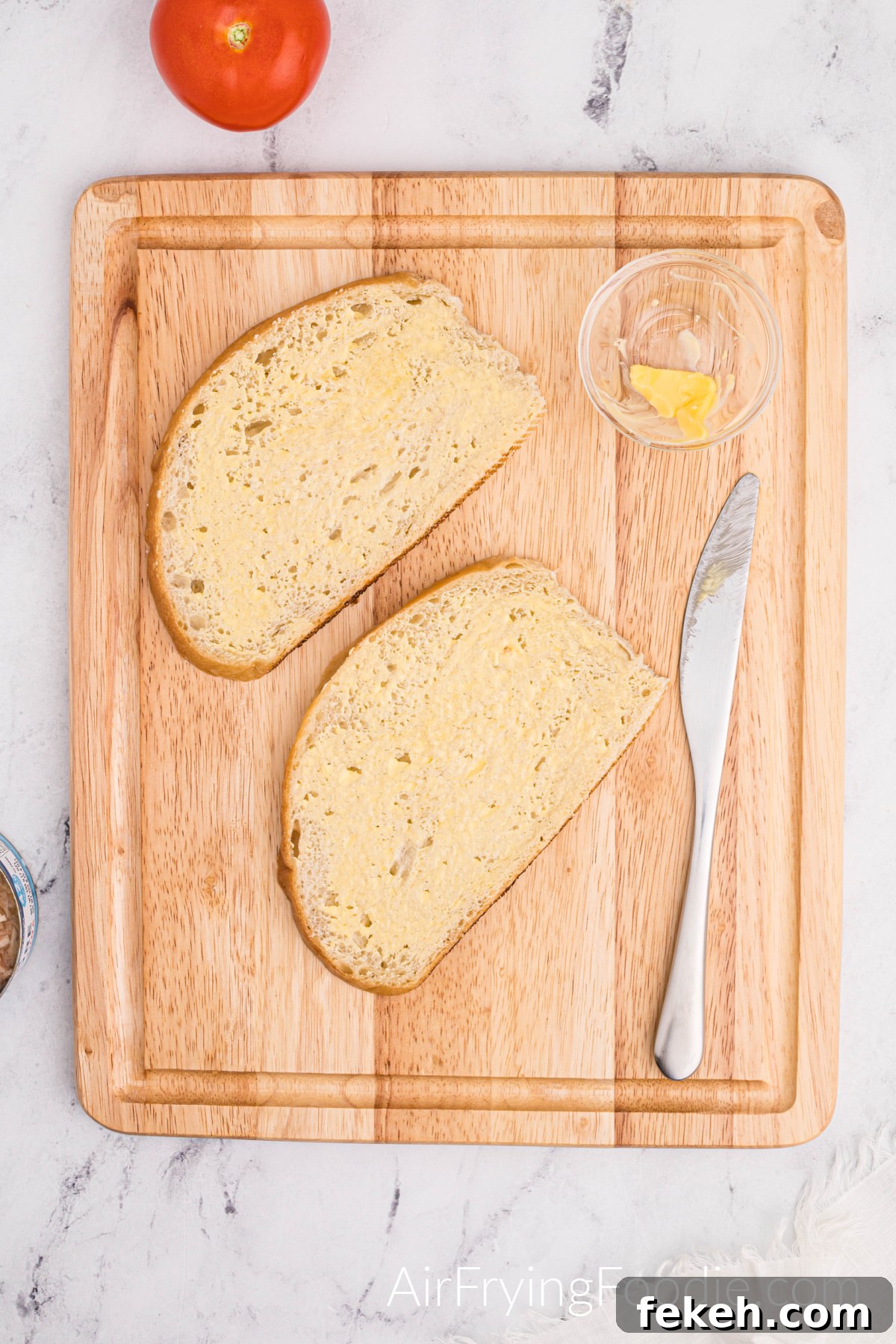 Crispy Air Fryer Tuna Melt Sandwich Recipe 4 Two slices of Texas toast buttered on a cutting board with a small glass bowl of butter and a butter knife. There is a tomato to the top left of the cutting board.
