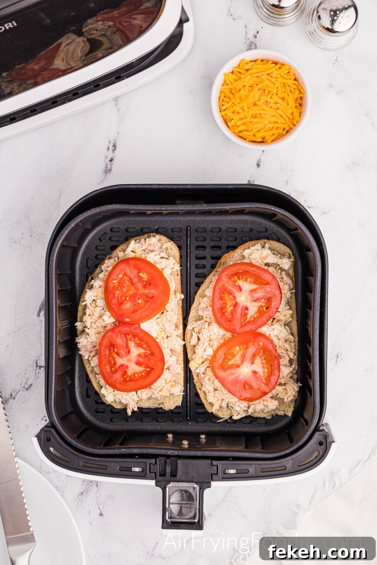 Crispy Air Fryer Tuna Melt Sandwich Recipe 8 Two slices of Texas toast in the air fryer basket topped with tuna and two slices of tomato on each piece of Texas toast. There is a small bowl of shredded cheese, a salt, and pepper shaker to the top right of the air fryer basket, and a small portion of the air fryer base if at the top left.