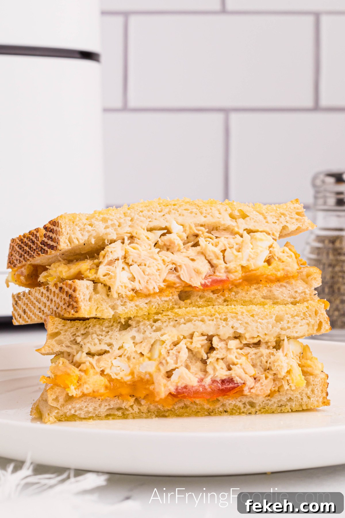 Crispy Air Fryer Tuna Melt Sandwich Recipe 10 Tuna melt sandwich on golden Texas toast cooked in the air fryer with tomato and cheese cut in half and served on a round white plate. There is a small portion of a pepper shaker to the right of the sandwich and a small part of the air fryer base to the left of the picture.