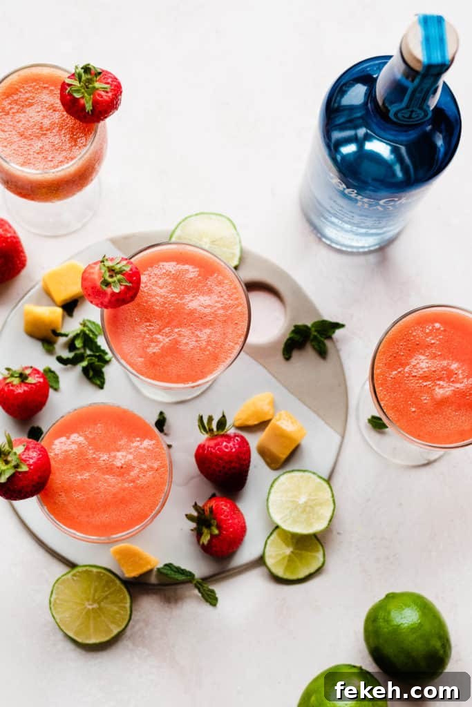 Three cocktail glasses filled with a bright pinky orange rum punch, with mangos, strawberries, and lime slices scattered around, and a blue rum bottle