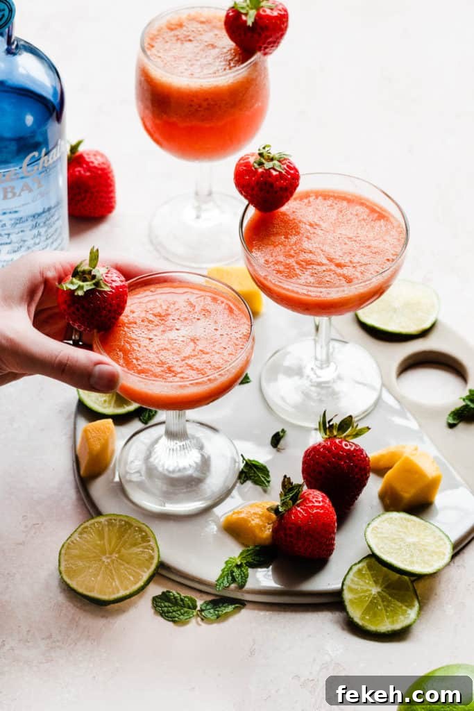 Three cocktail glasses filled with a bright pinky orange frozen punch, and garnished with fresh strawberries, with fruit scattered around, and a hand picking up one of the glasses