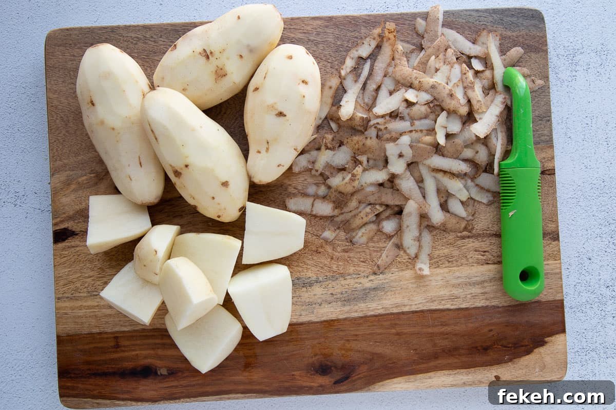 peeled whole potatoes and peeled chopped potatoes on a cutting board with potato peels and a peeler.