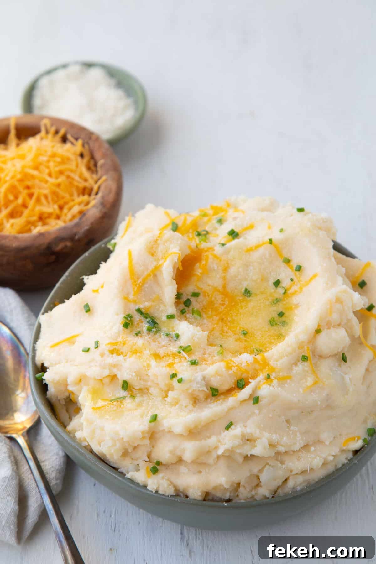 cheesy mashed potatoes topped with chives and butter in a green bowl, next to smaller bowls of cheddar and parmesan.