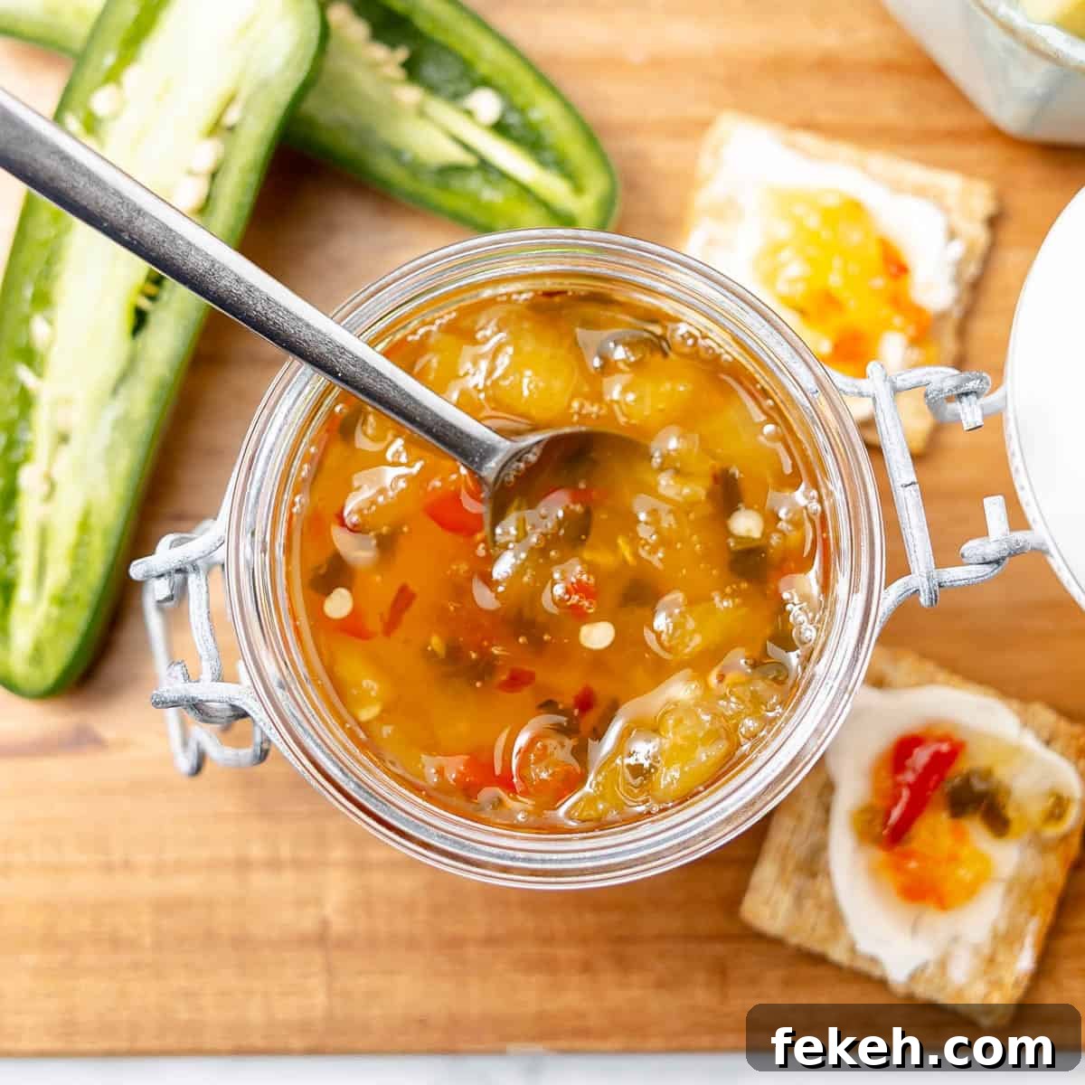Pineapple Jalapeño Jam Recipe: Sweet-Spicy Preserves Guide 14 An open jar of pepper jelly with a spoon inside, surrounded by sliced jalapeños and crackers topped with cream cheese and jelly on a wooden surface.