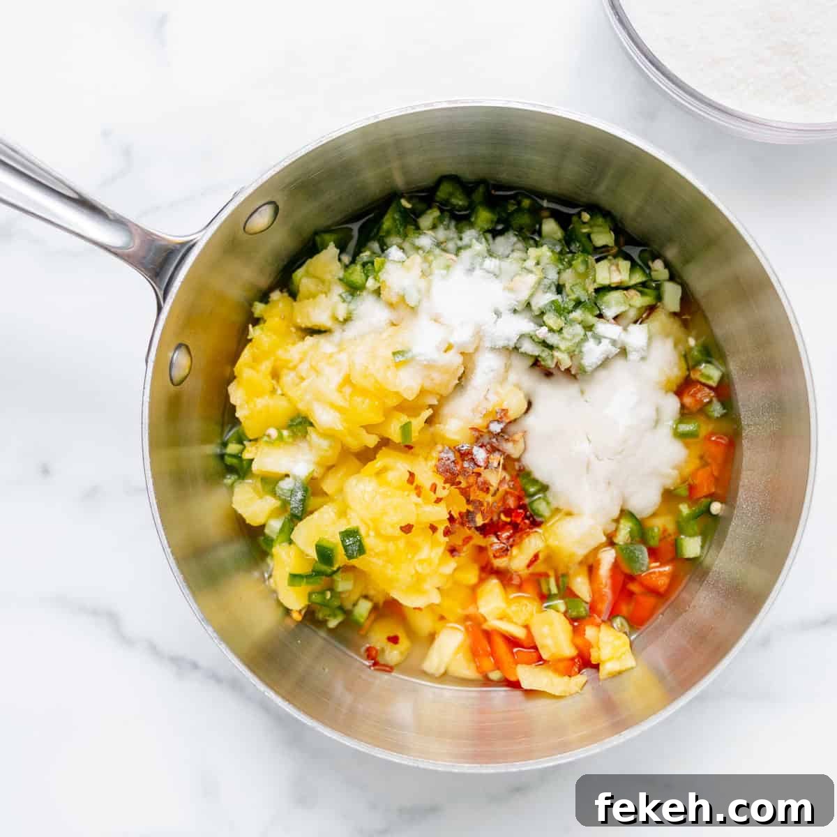 Pineapple Jalapeño Jam Recipe: Sweet-Spicy Preserves Guide 5 A saucepan containing chopped vegetables, crushed pineapple, sugar, red pepper flakes, and other ingredients on a white marble surface.