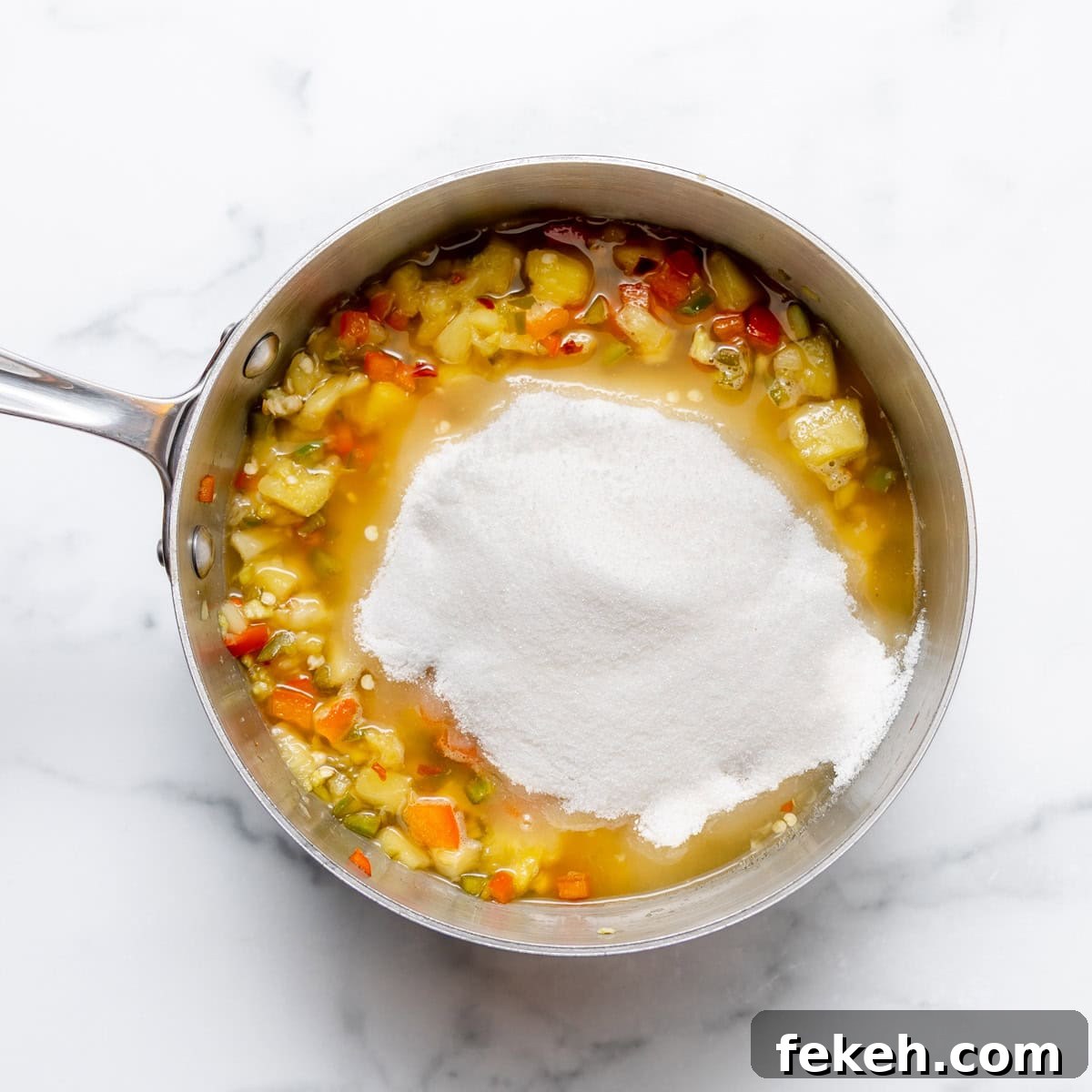 Pineapple Jalapeño Jam Recipe: Sweet-Spicy Preserves Guide 6 A saucepan containing chopped vegetables and liquid on a white surface, with a mound of white granulated sugar added on top.