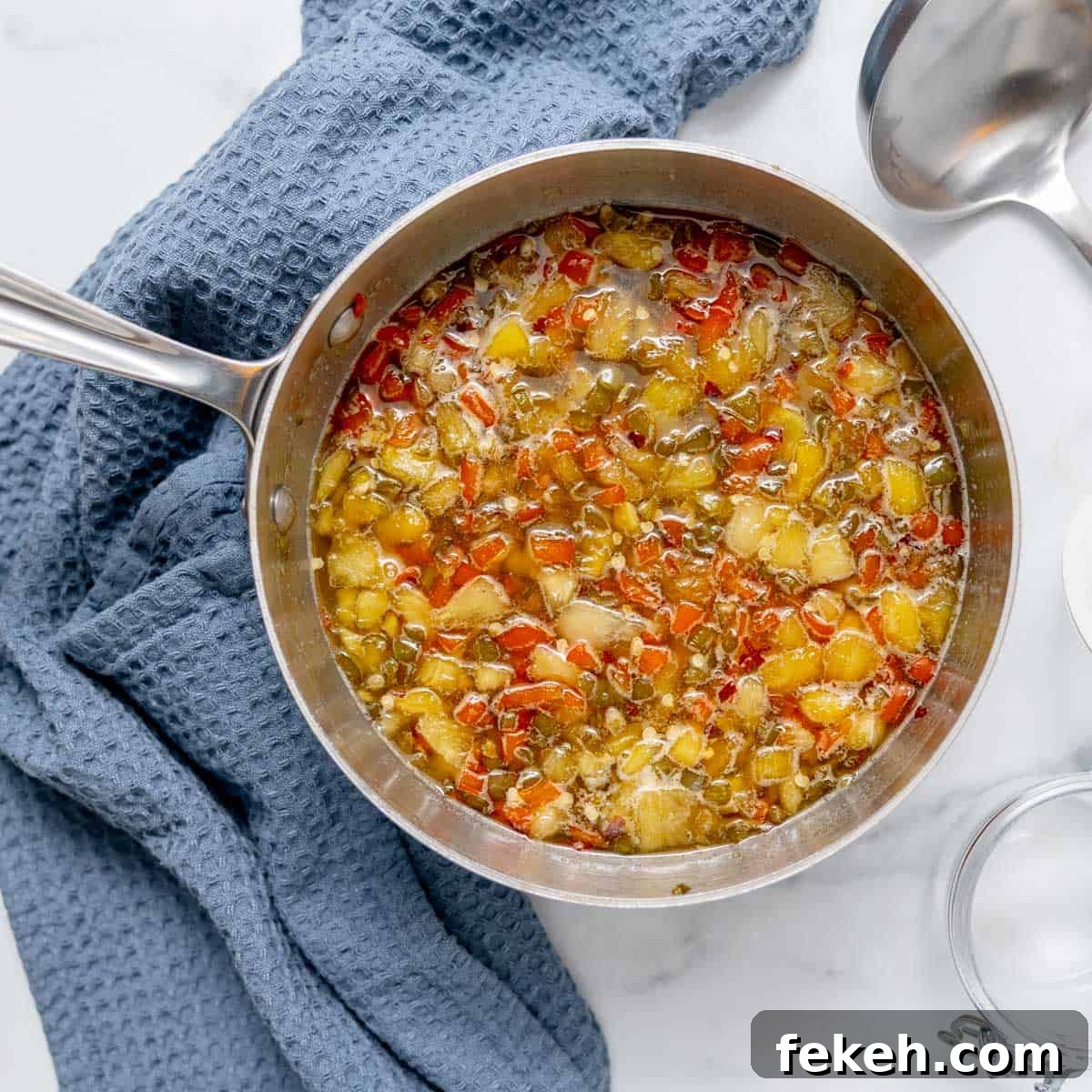 Pineapple Jalapeño Jam Recipe: Sweet-Spicy Preserves Guide 7 A saucepan filled with a chunky vegetable mixture sits on a white surface next to a blue textured cloth and a metal spoon.