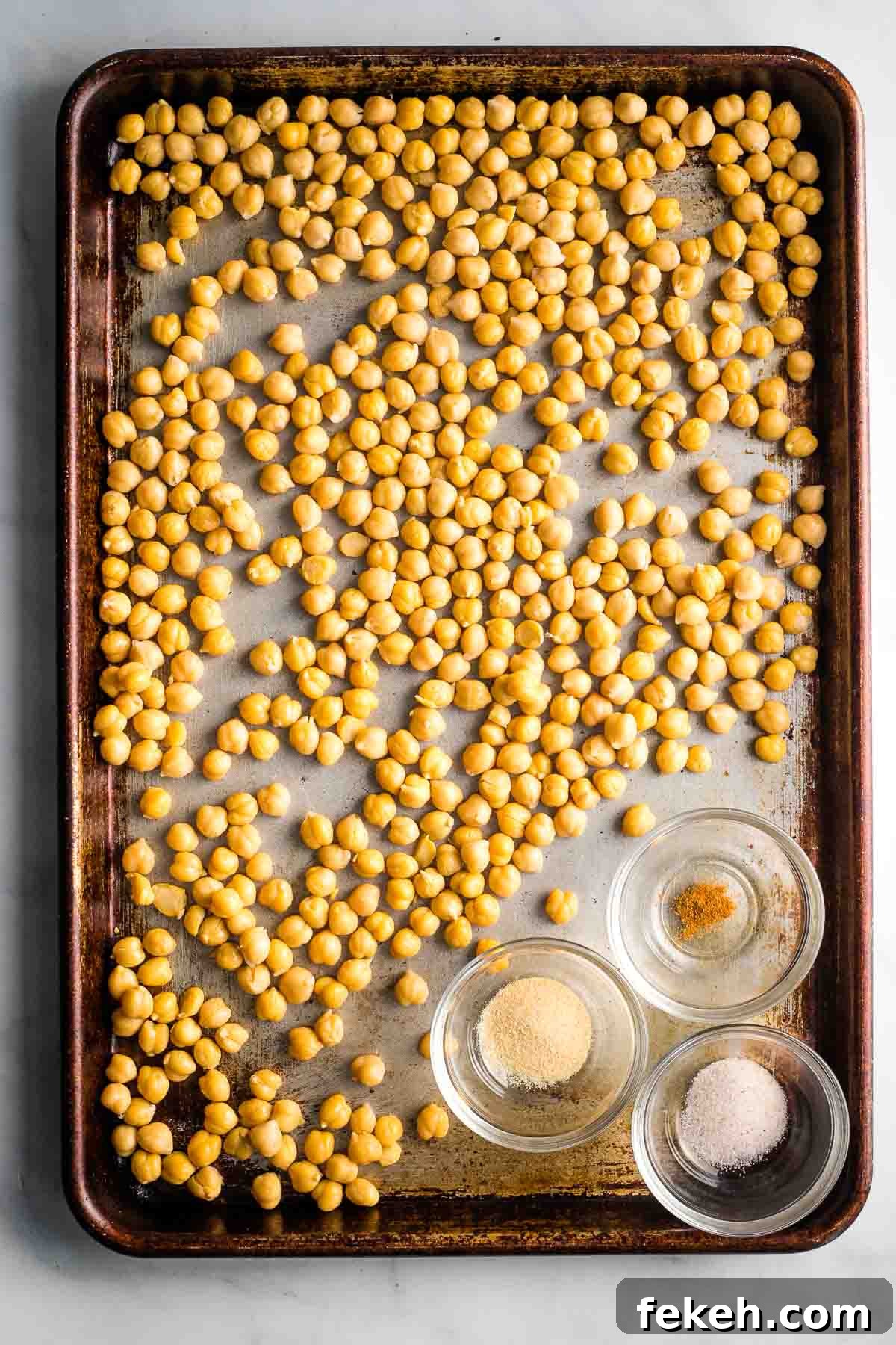 Chickpeas in a single layer on a baking sheet with three small dishes with spices.
