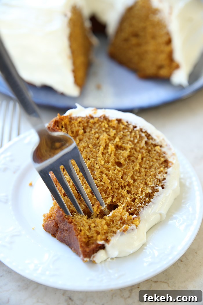 A slice of pumpkin bundt cake with cream cheese frosting on a dessert plate with a fork inserted into the cake and faded into the background is a pumpkin bundt cake with cream cheese frosting with a piece cut out on a round plate.