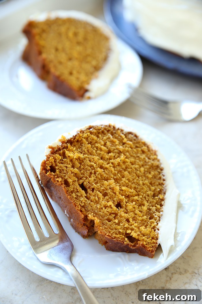A slice of pumpkin bundt cake with cream cheese frosting on a dessert plate with a fork next to it. Faded in the background is another piece of cake on a small plate and a part of a pumpkin bundt cake on a blue round plate.