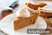 A slice of pumpkin pie with a dollop of whipping cream on top sitting on a small dessert plate. Faded in the background is a pie server and a pumpkin pie with a slice cut out of it.