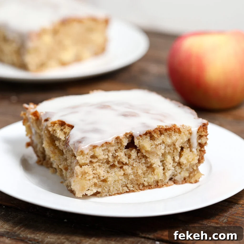 44 Gluten-Free Dairy-Free Cake Recipes for Every Occasion 16 A square slice of glazed Apple Fritter Cake sits on a white plate, with a whole apple and another plate of cake blurred in the background on a wooden table.