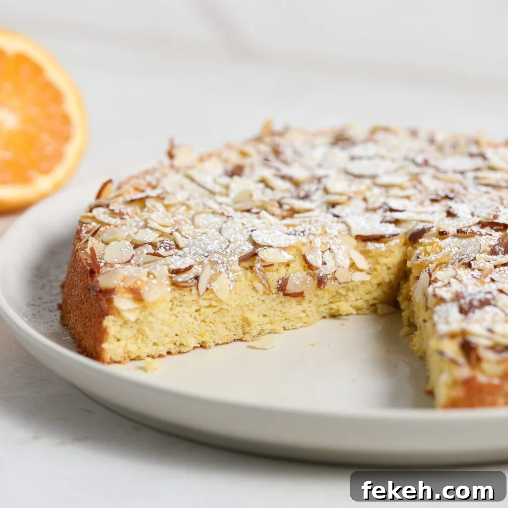 44 Gluten-Free Dairy-Free Cake Recipes for Every Occasion 18 A round Orange Almond Cake topped with sliced almonds and powdered sugar sits on a white plate. A slice has been removed, revealing its moist texture, and a halved orange is visible in the background.