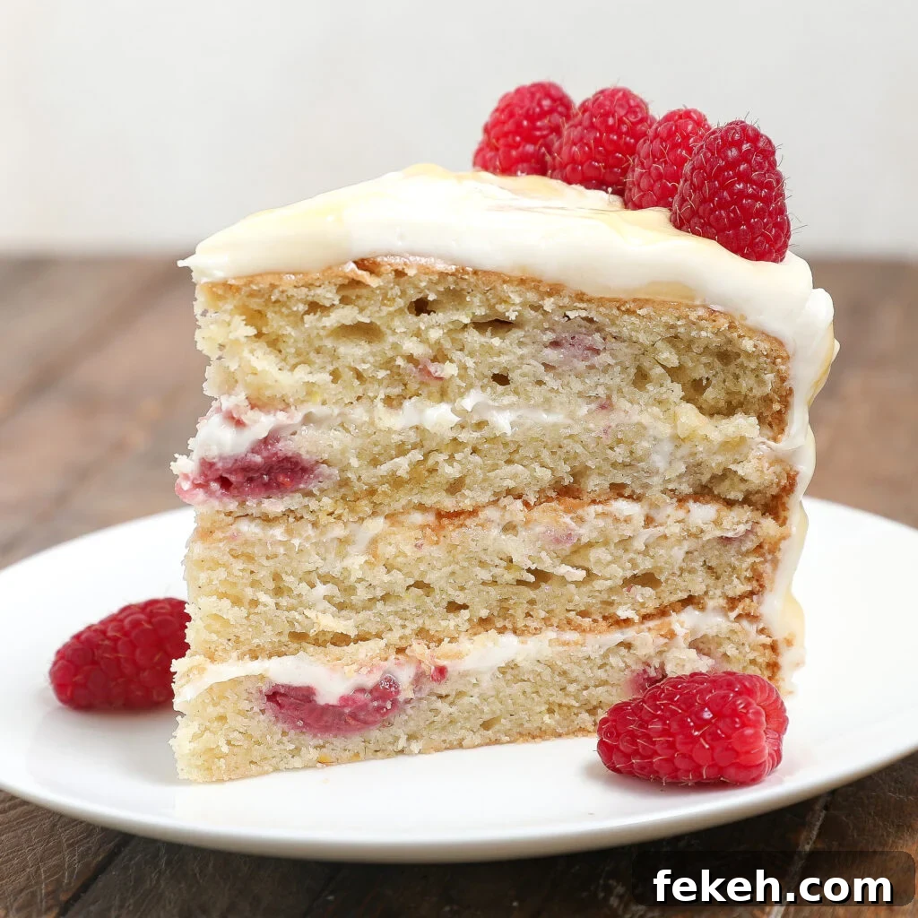 44 Gluten-Free Dairy-Free Cake Recipes for Every Occasion 4 A three-layer slice of Raspberry Lemon Cake with raspberries and cream filling, topped with frosting and fresh raspberries, sits on a white plate on a wooden surface.