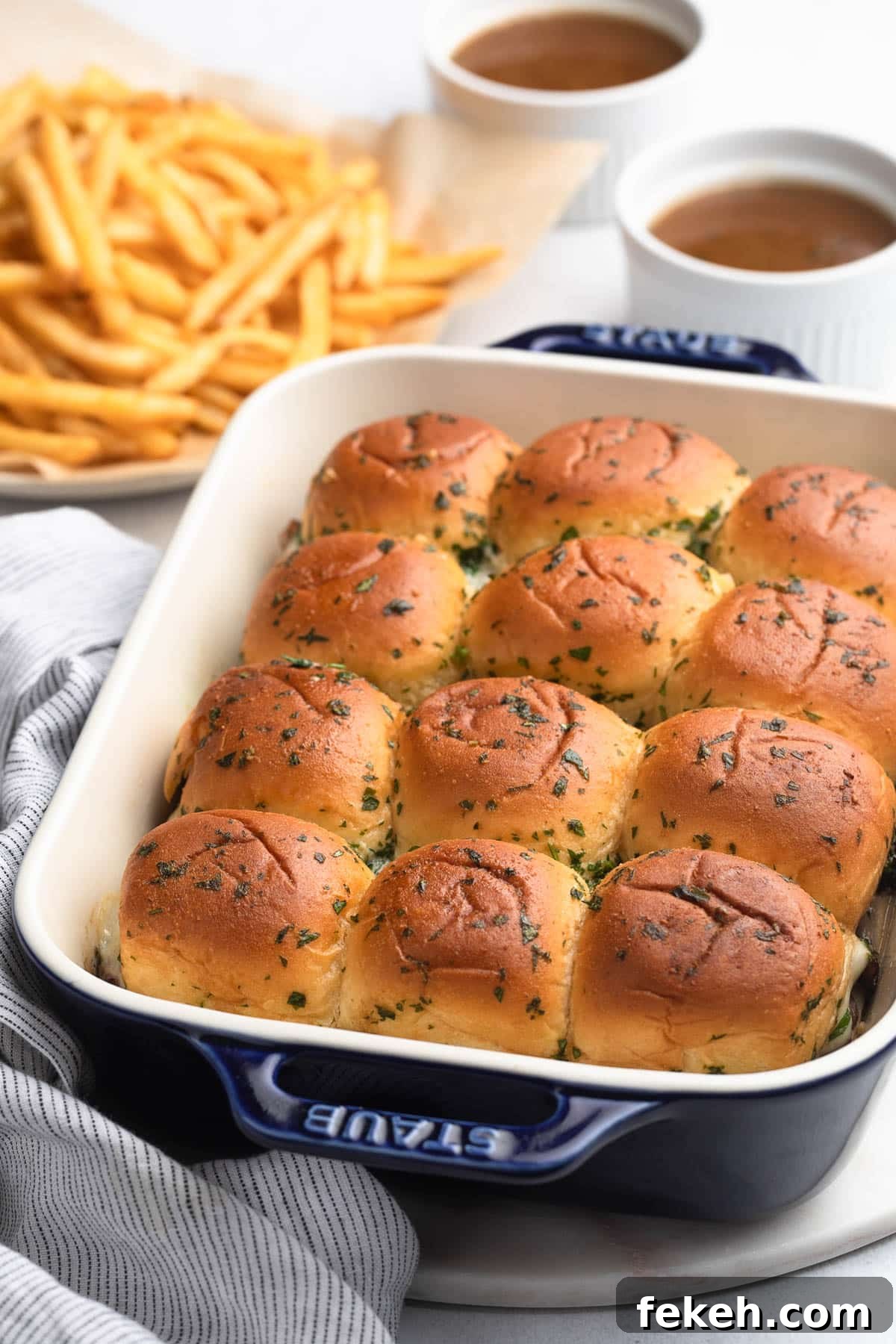 baked french dip sliders in a white and blue baking dish with bowls of au jus and a plate of french fries