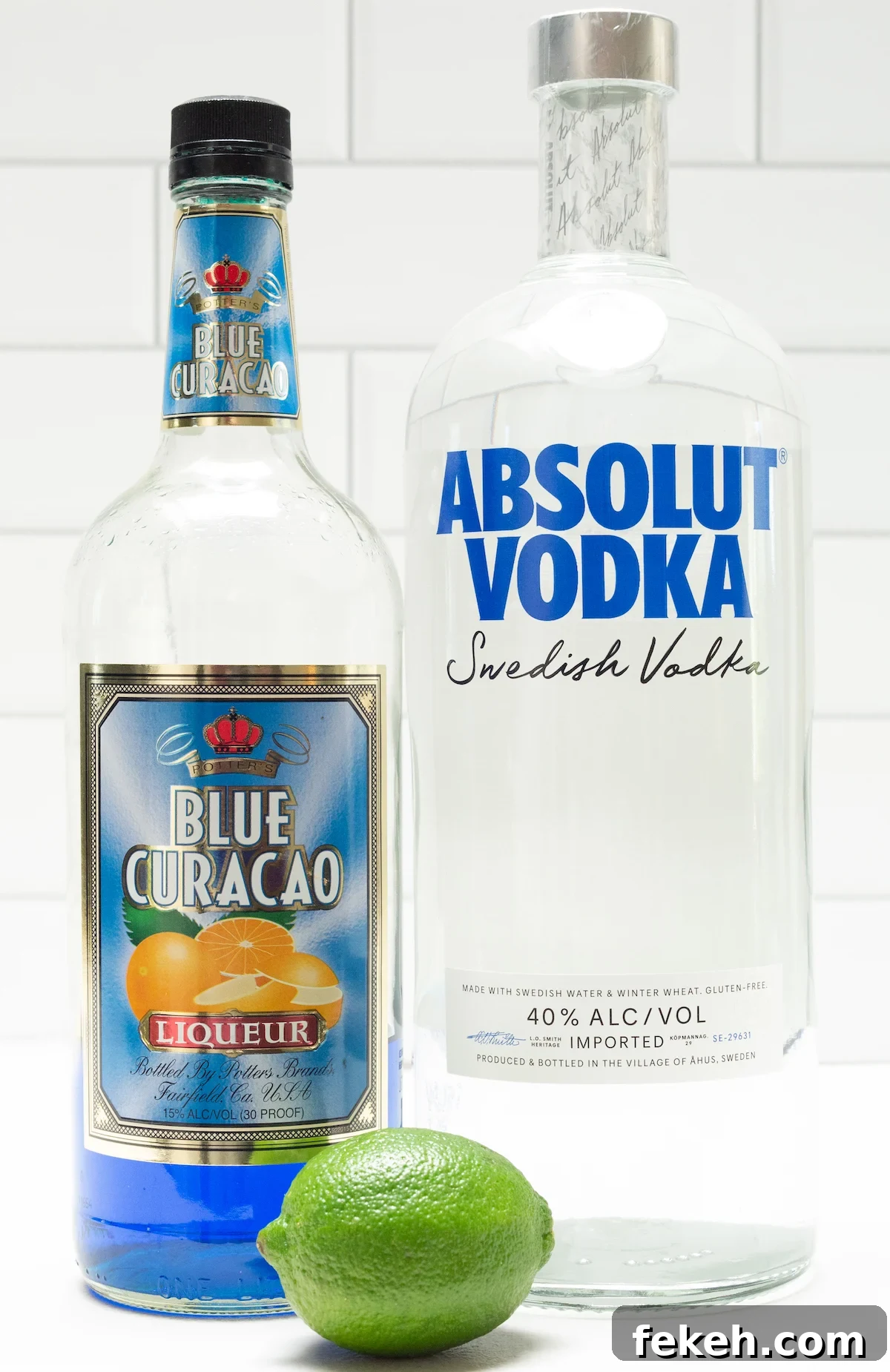 Blue Kamikaze Cocktail Recipe: How to Make Vibrant Shooter Shots 3 A bottle of blue curacao and a bottle of vodka in front of a white tile background.