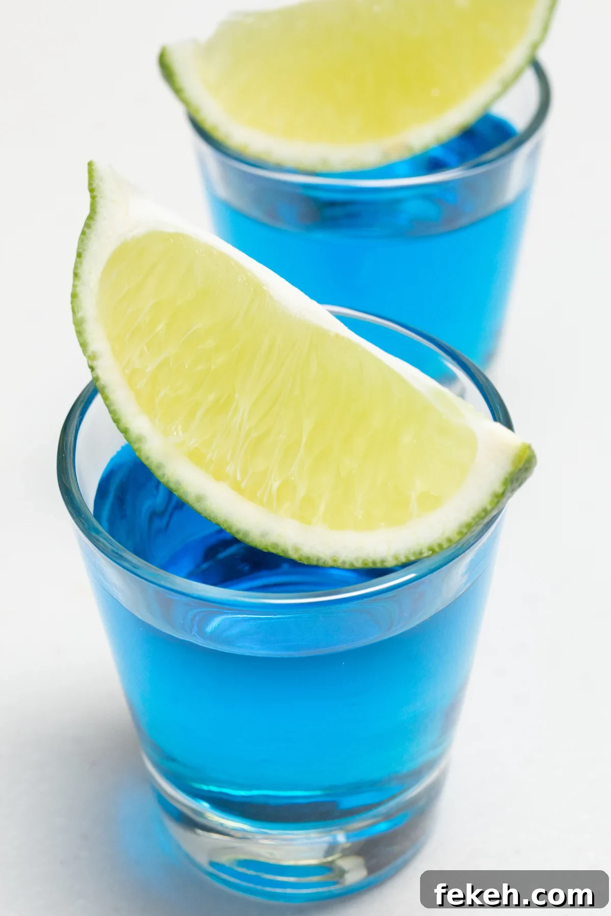 Blue Kamikaze Cocktail Recipe: How to Make Vibrant Shooter Shots 4 Two shot glasses filled with bright blue liquid are on a white background. Fresh cut lime wedges sit across the top of each shot glass.