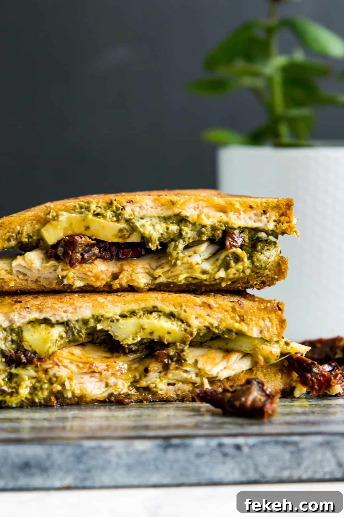 Grilled Turkey and Pesto Sandwich Recipe 7 A sandwich with turkey, sun dried tomatoes, mozzarella cheese and pesto in it that has been cut in half and stacked on a cutting board.