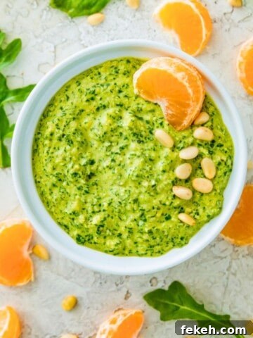A white bowl filled with arugula pesto and garnished with pine nuts and an orange slice.