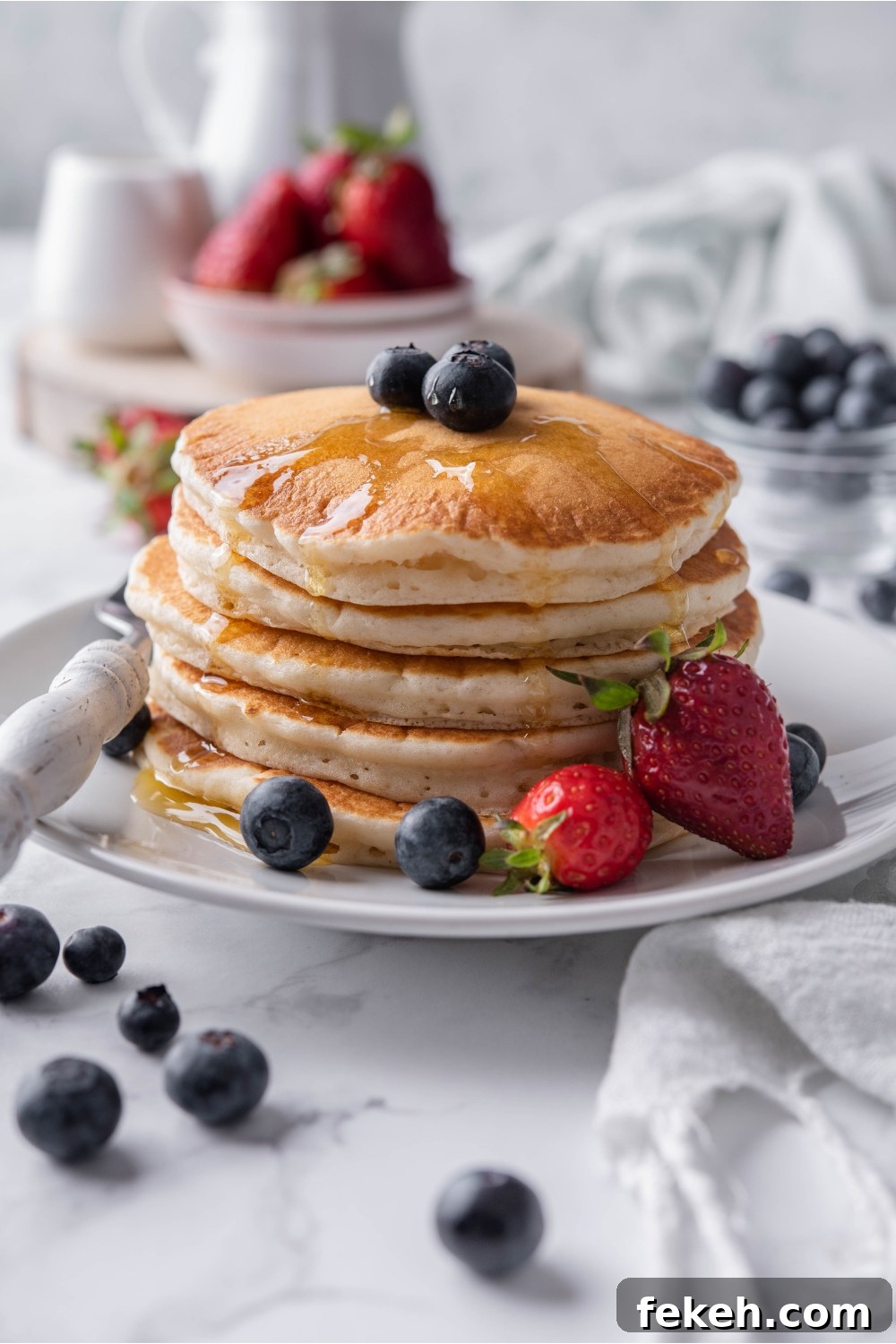 Light and Fluffy Vegan Pancakes Without Eggs 2 A stack of pancakes topped with maple syrup and blueberries on a white plate with a fork and strawberries on the plate beside the pancakes. Berries are scattered on the counter and in the background.