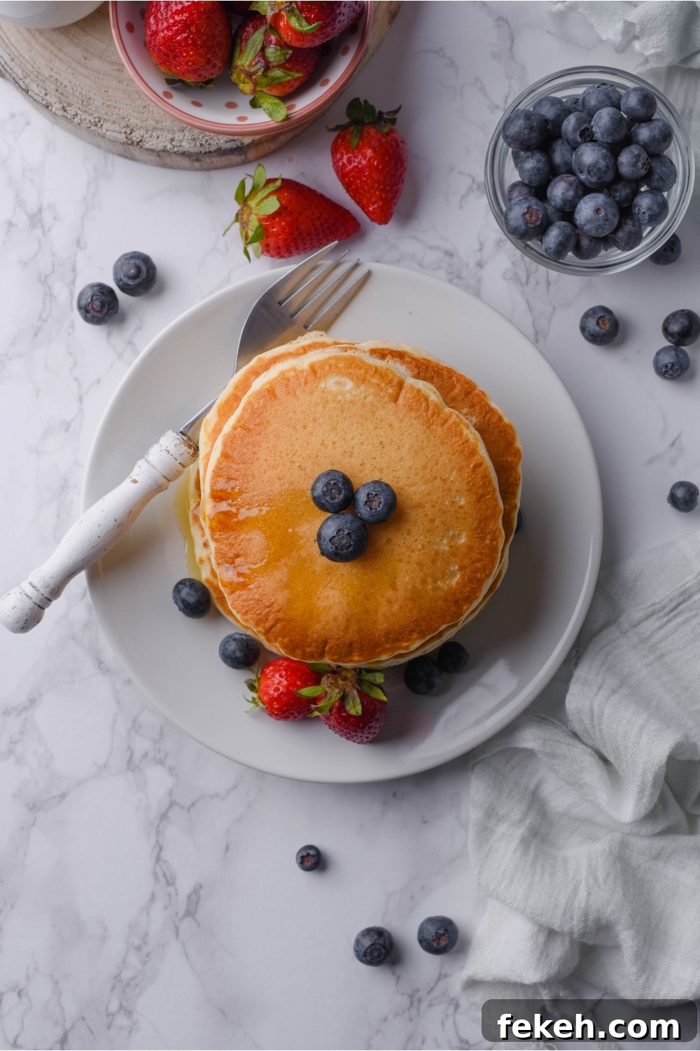 Light and Fluffy Vegan Pancakes Without Eggs 11 Overhead view of a stack of pancakes topped with maple syrup and blueberries. Surrounding the pancakes are blueberries and strawberries scattered on the countertop.