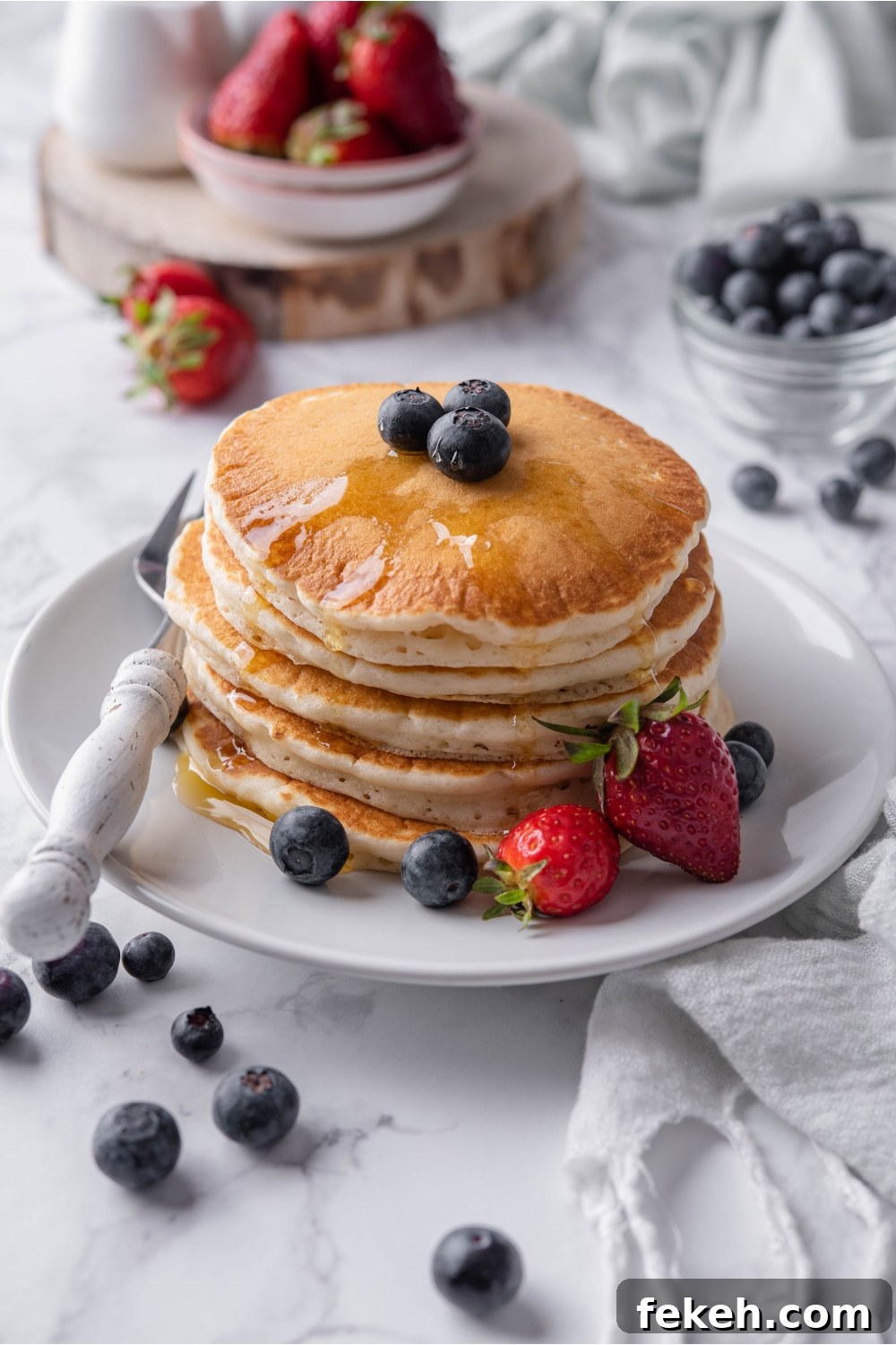 Light and Fluffy Vegan Pancakes Without Eggs 12 A stack of pancakes topped with maple syrup and blueberries on a white plate with a fork and strawberries on the plate beside the pancakes. Berries are scattered on the counter and in the background.
