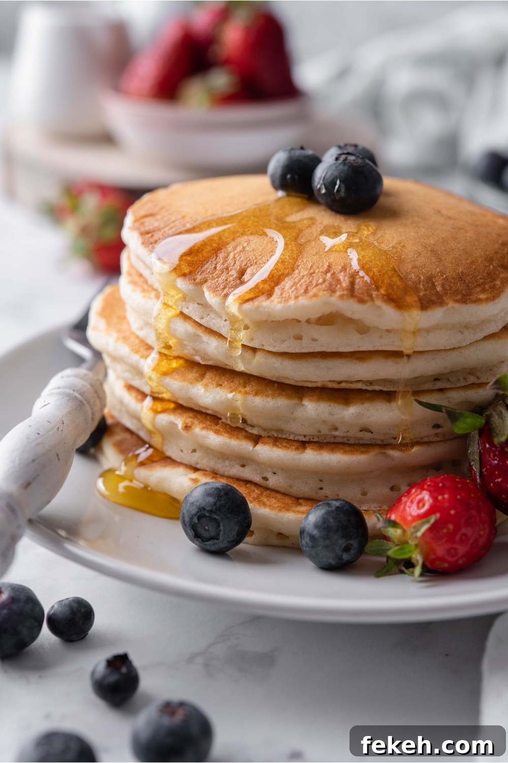 Light and Fluffy Vegan Pancakes Without Eggs 13 A stack of pancakes topped with maple syrup and blueberries on a white plate with a fork and strawberries on the plate beside the pancakes. Berries are scattered on the counter and in the background.