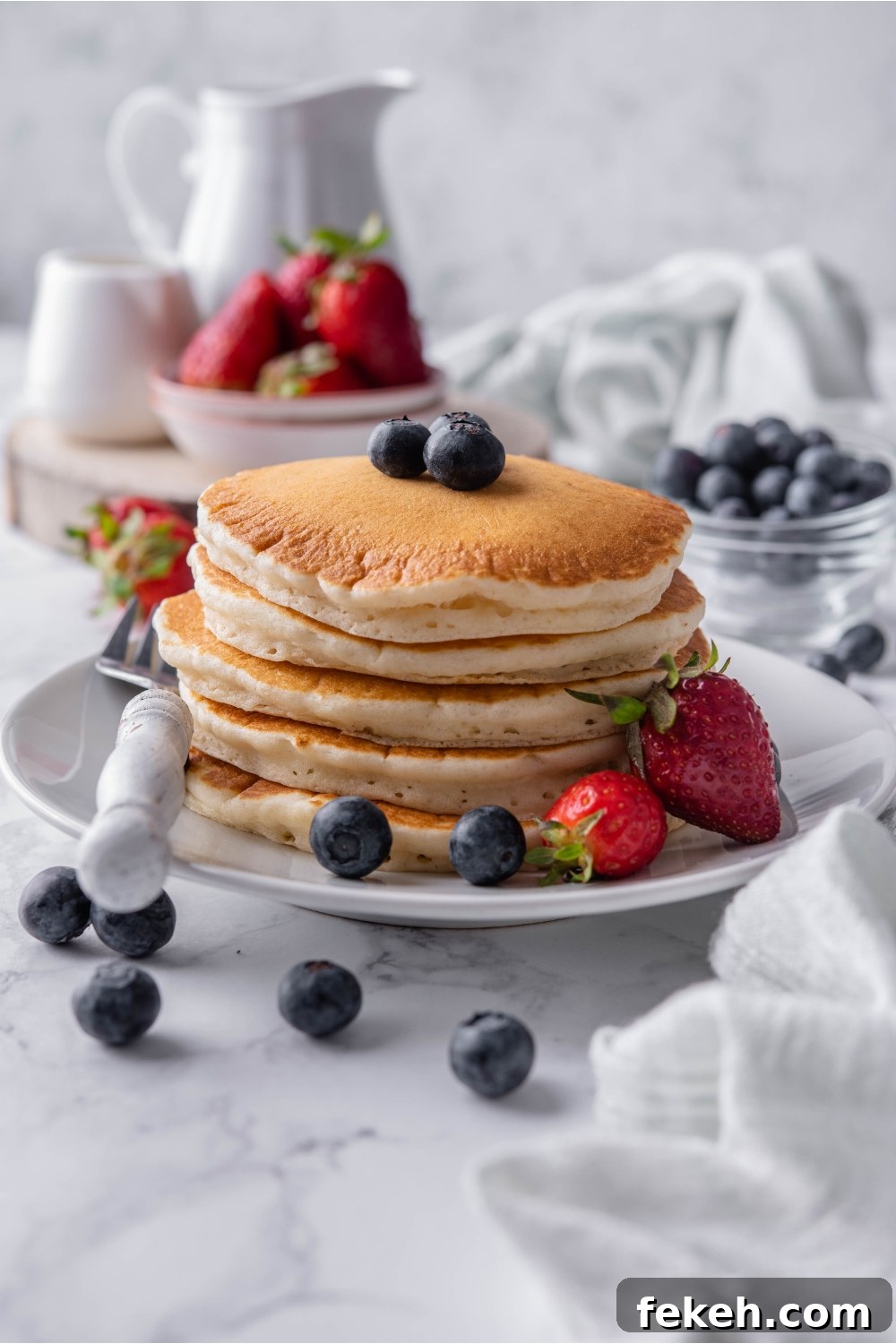 Light and Fluffy Vegan Pancakes Without Eggs 9 A stack of pancakes topped with blueberries on a white plate with a fork and strawberries on the plate beside the pancakes. Berries are scattered on the counter and in the background.
