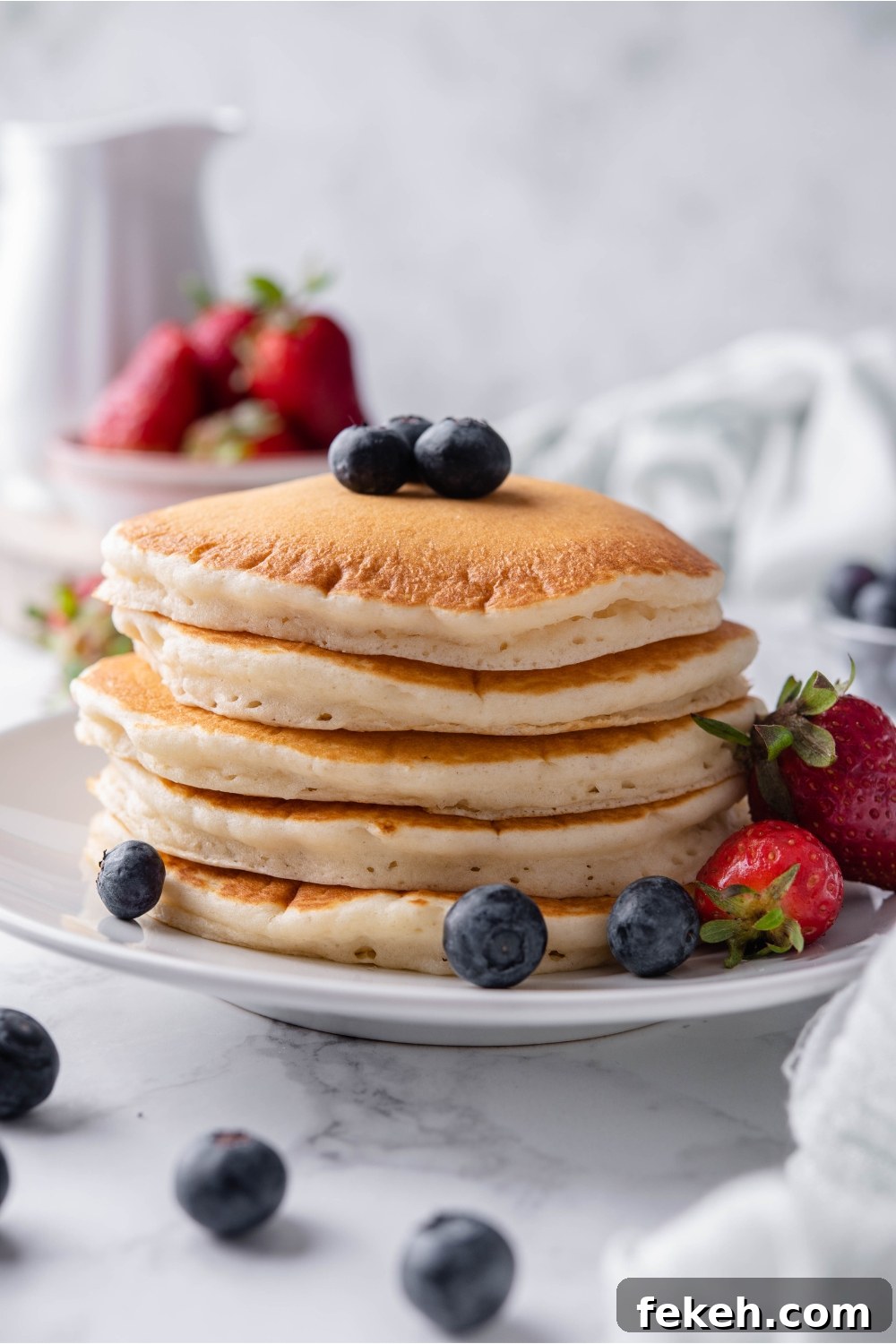 Light and Fluffy Vegan Pancakes Without Eggs 10 A stack of pancakes topped with blueberries on a white plate with a fork and strawberries on the plate beside the pancakes. Berries are scattered on the counter and in the background.