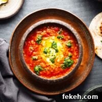 Spicy Harissa Lentil Soup with Fresh Cilantro Salsa 7 A brown ceramic bowl with bright red harissa lentil soup with a scoop of yoghurt and dots of green salsa. The bowl is on a dark surface with pieces of flatbread visible on the side.