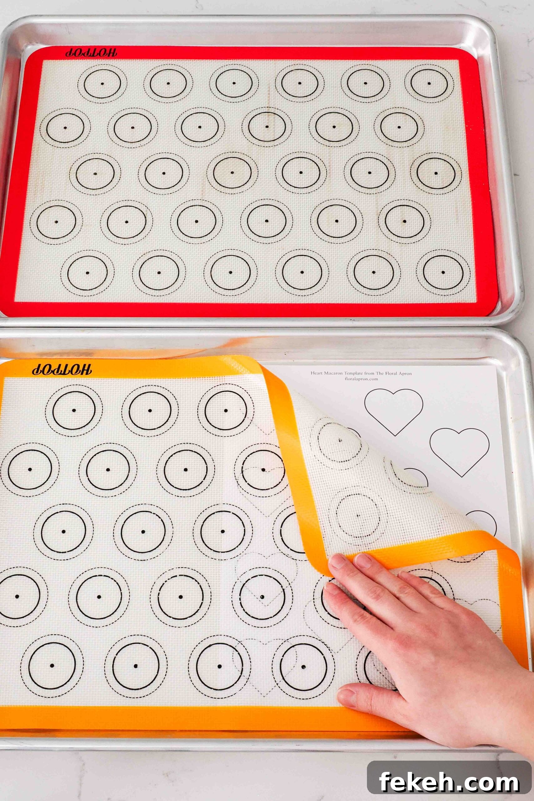 Two baking sheets with silicone macaron mats and a paper template with hearts underneath.
