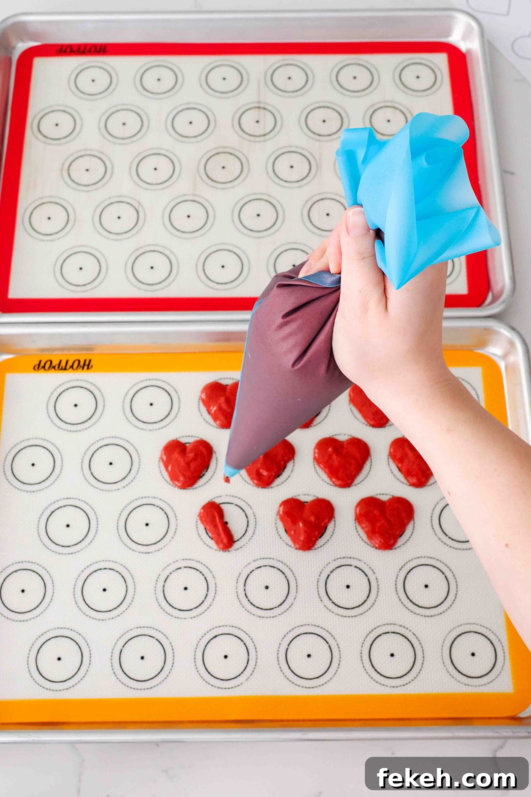 A hand pipes the left half of a heart-shaped macaron on silicone macaron mats.