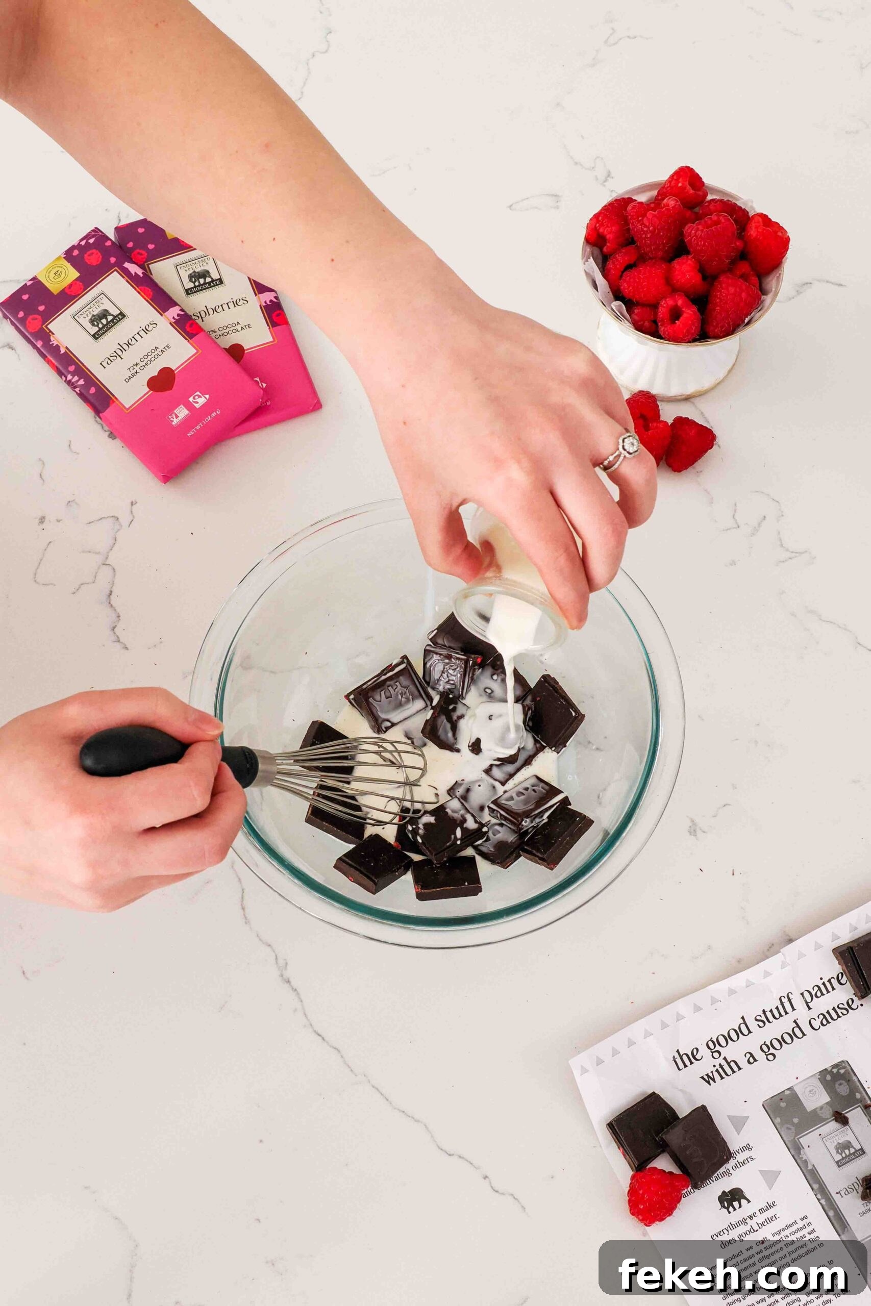 A hand pours heavy cream into a bowl of pieces of raspberry chocolate.