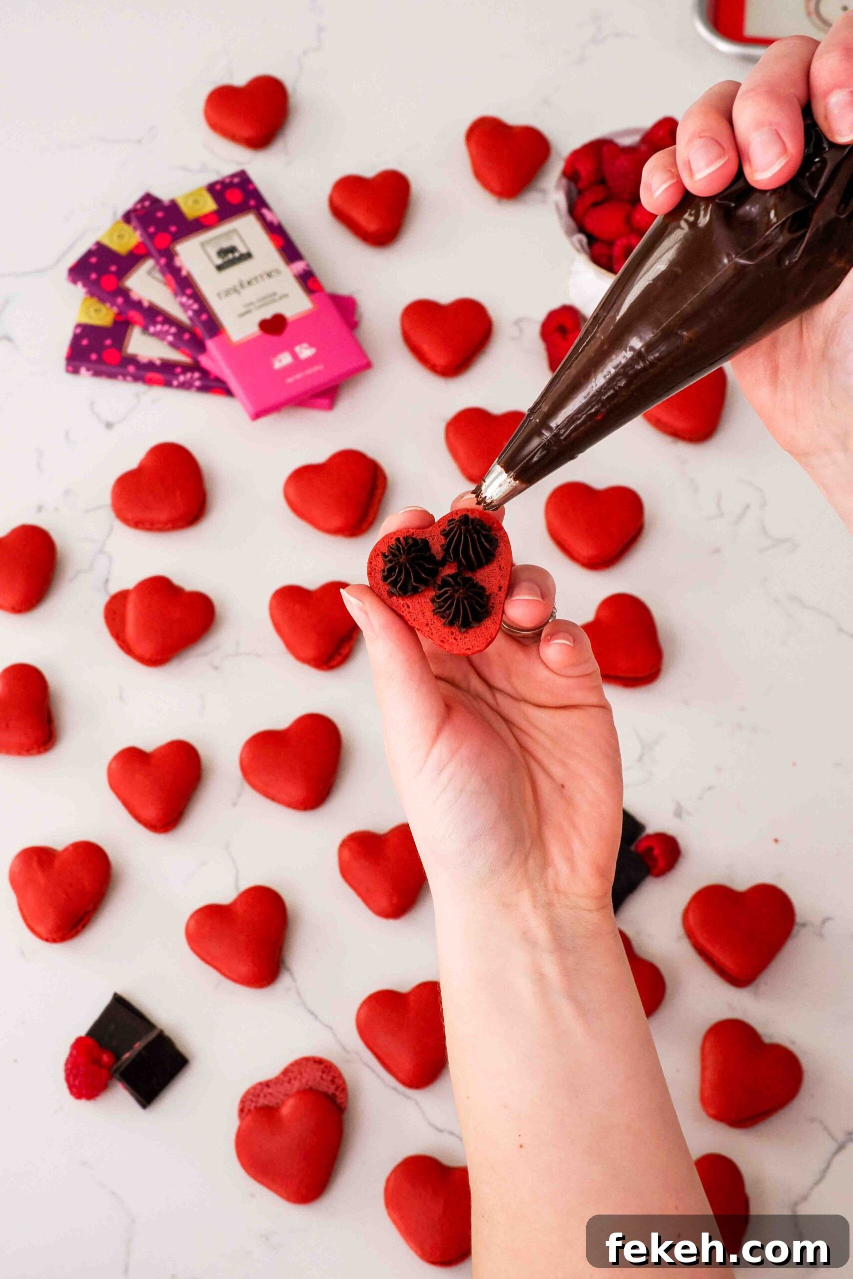 A hand pipes three dots of raspberry chocolate ganache onto the underside of a heart-shaped macaron shell.