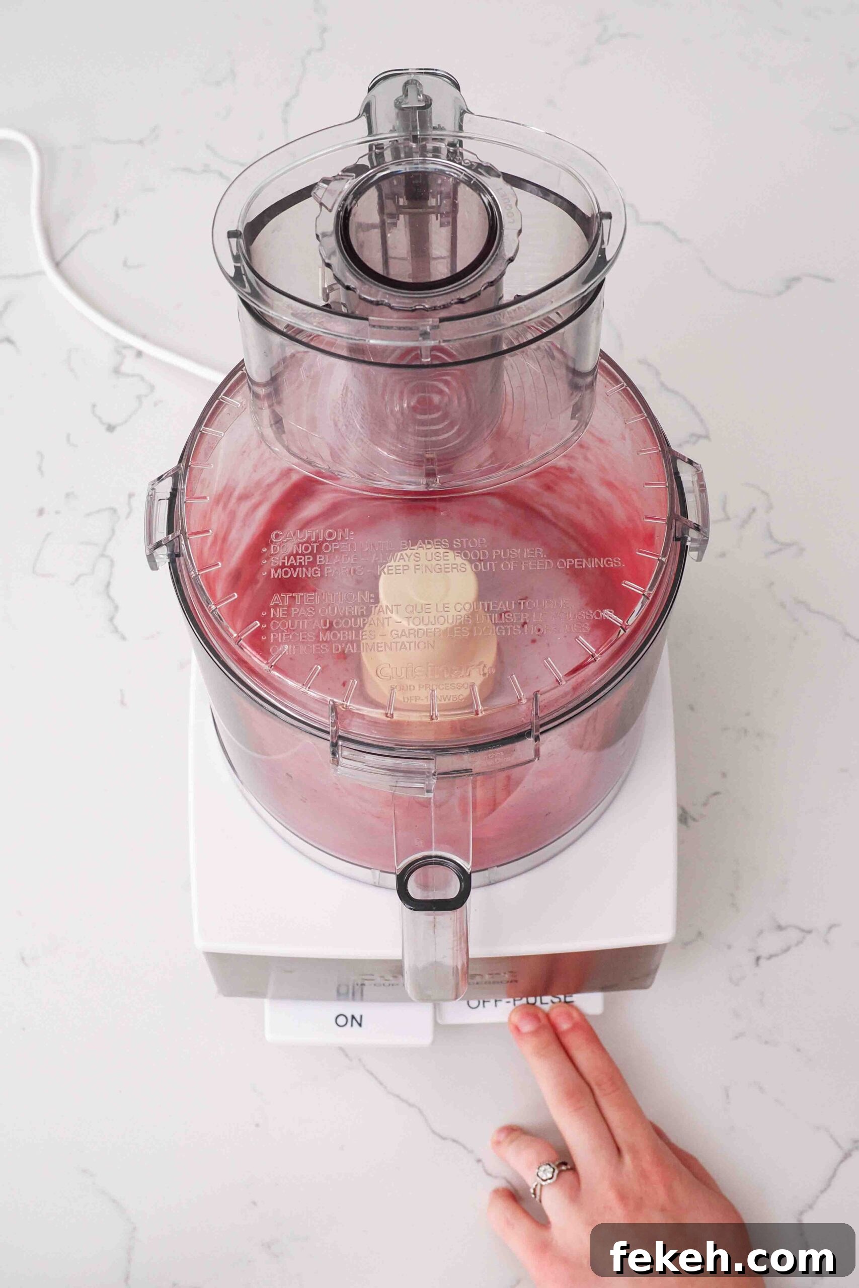 A hand presses the "pulse" button on a food processor with freeze-dried raspberries.