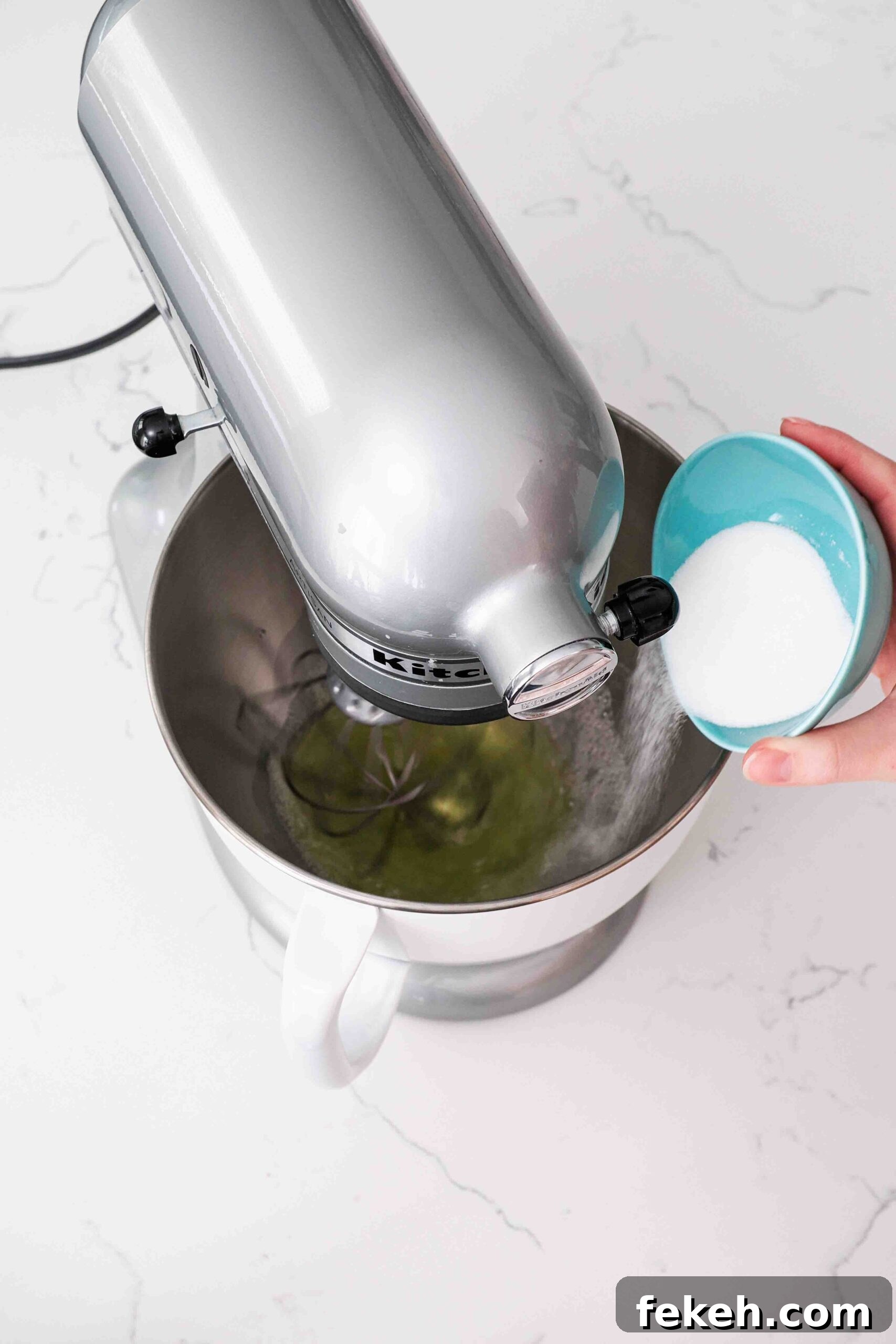 A hand sprinkles granulated sugar into the bowl of a stand mixer whisking egg whites.